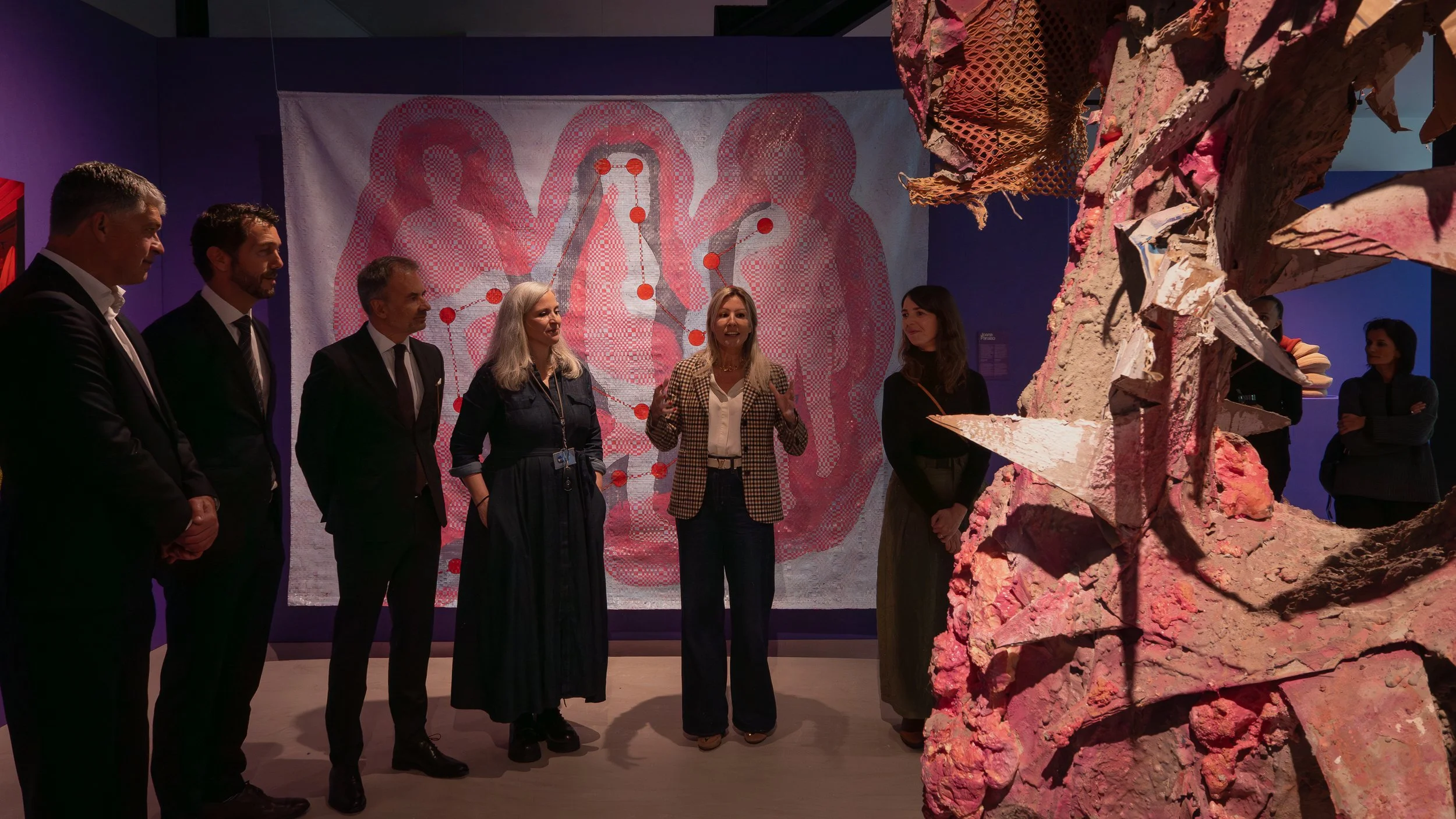 Grupo de pessoas observando uma escultura artística feita de rochas coloridas em uma galeria de arte, com fundo de uma obra de arte abstrata de fundo