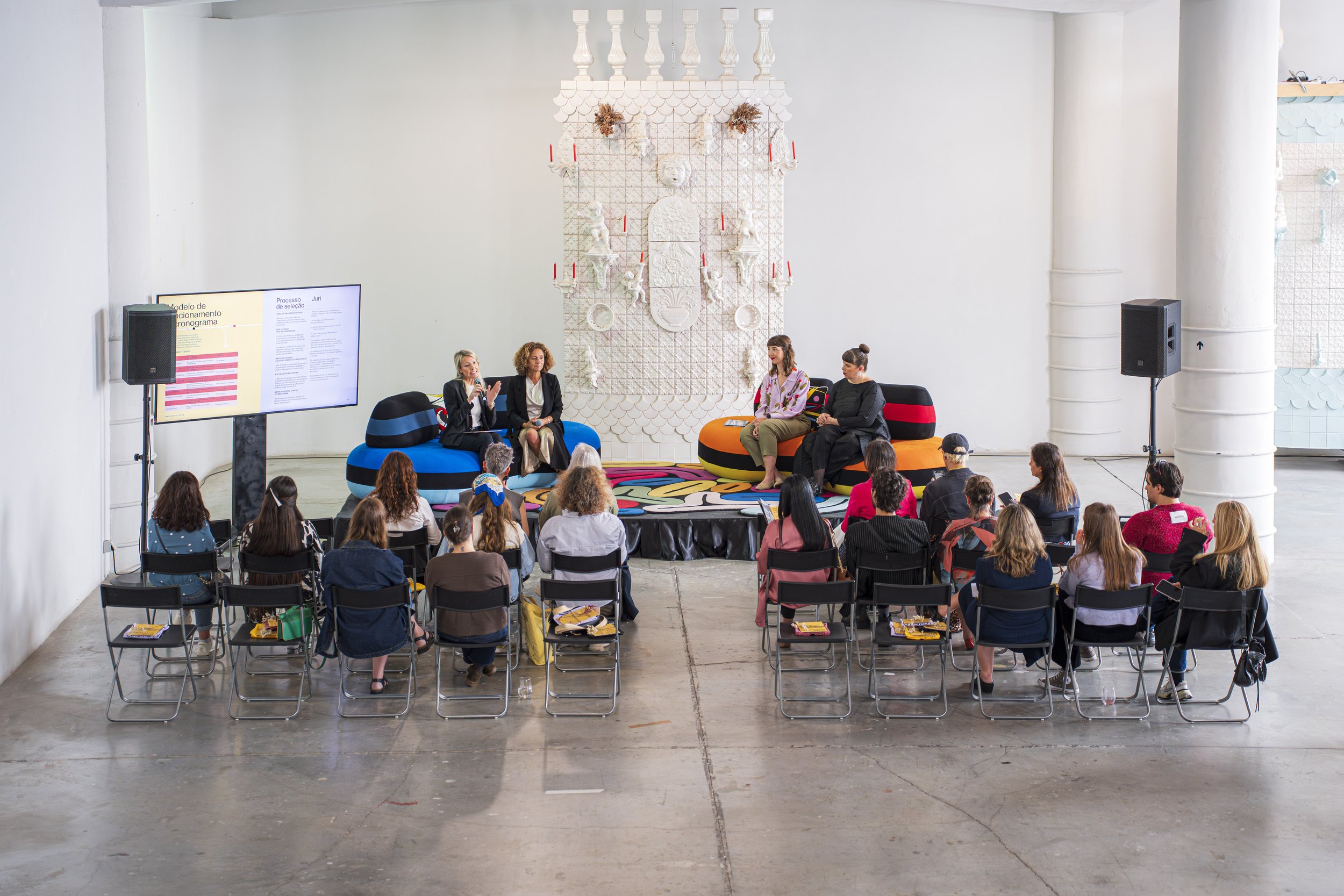 Painel de apresentação digital exibindo texto em português, com pessoas sentadas assistindo a uma palestra ou painel de discussão com um fundo decorado com arte em relevo branca e velas vermelhas, em um espaço amplo com paredes brancas e pilares.