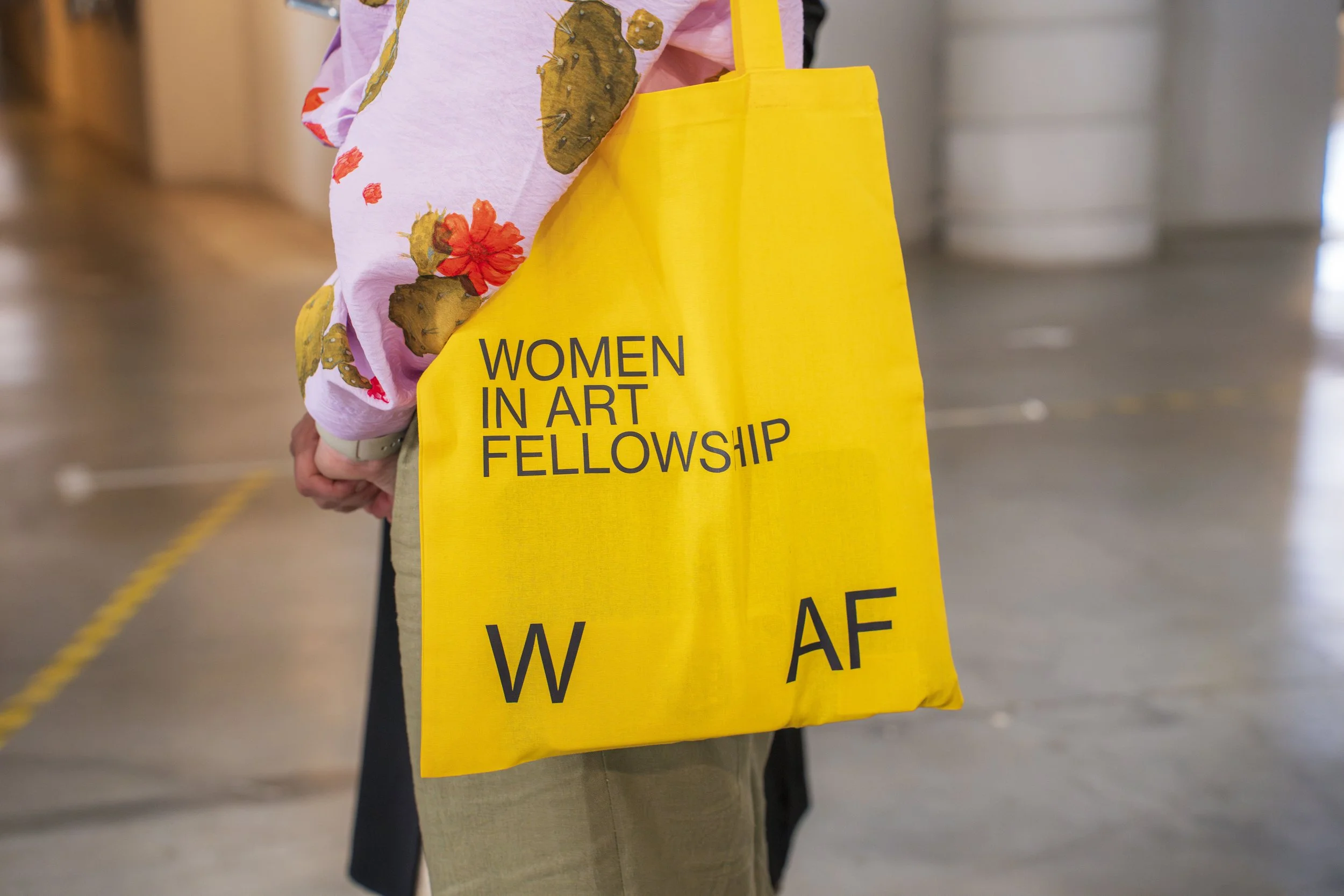 Pessoa segurando uma sacola amarela com a inscrição "Women in Art Fellowship", usando um casaco com estampa de borboletas e flores em fundo desfocado.