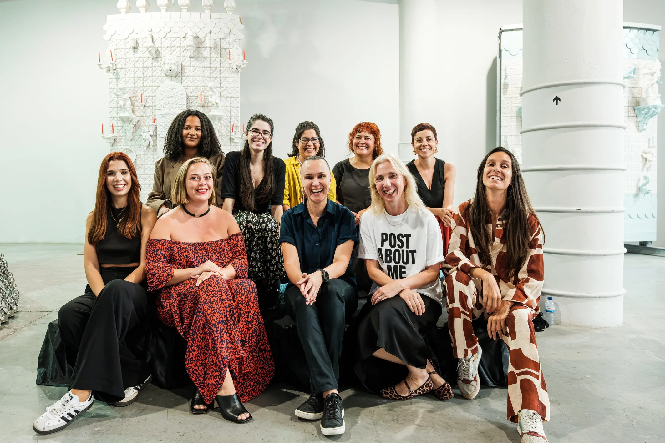 Grupo de pessoas sorrindo, posando para uma foto, em um ambiente interno com paredes brancas e decoração artística de fundo.