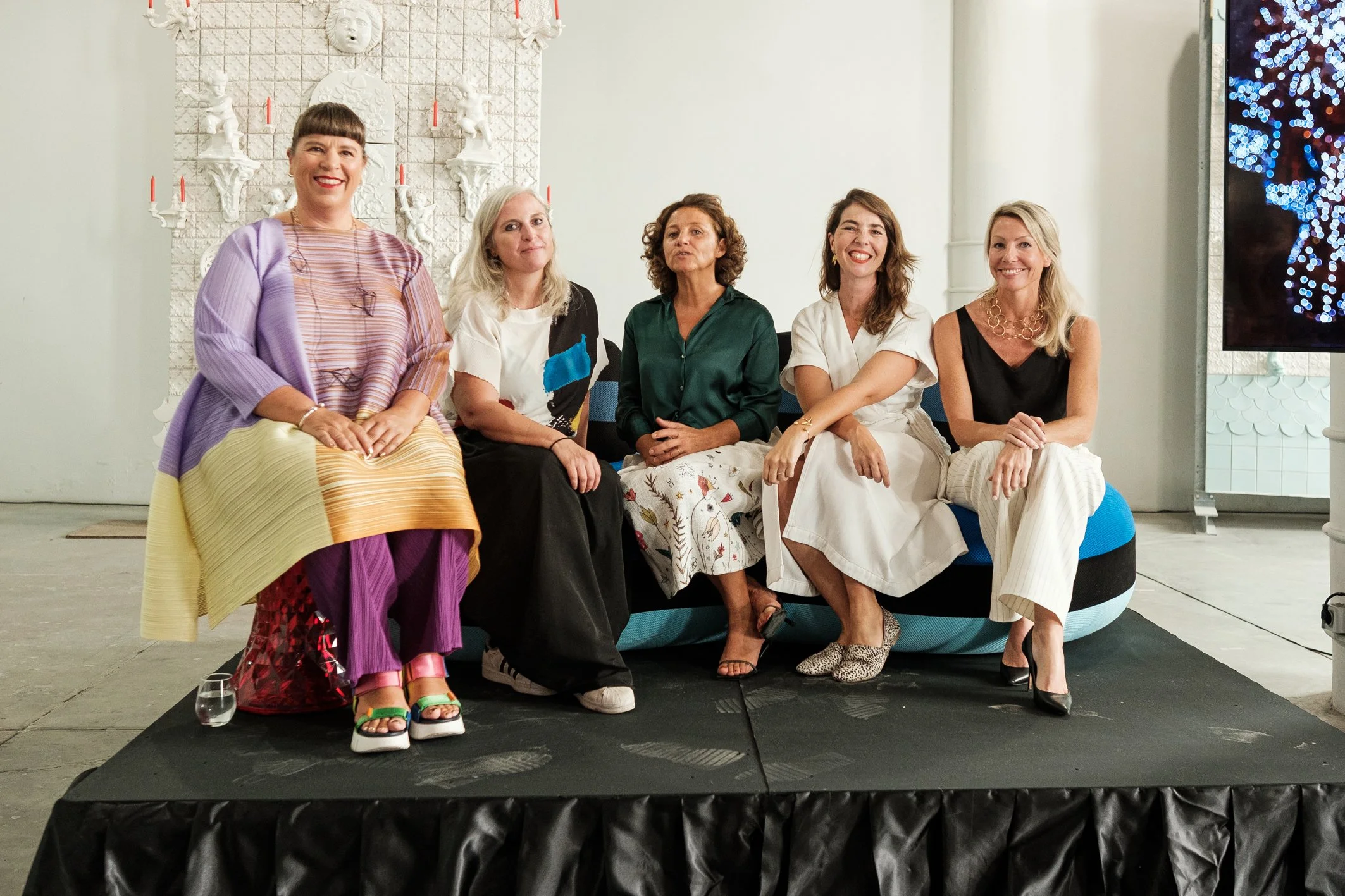 Grupo de cinco mulheres sentadas em uma plataforma, sorrindo para a câmera, com decoração branca ao fundo e uma tela à direita mostrando luzes brilhantes.