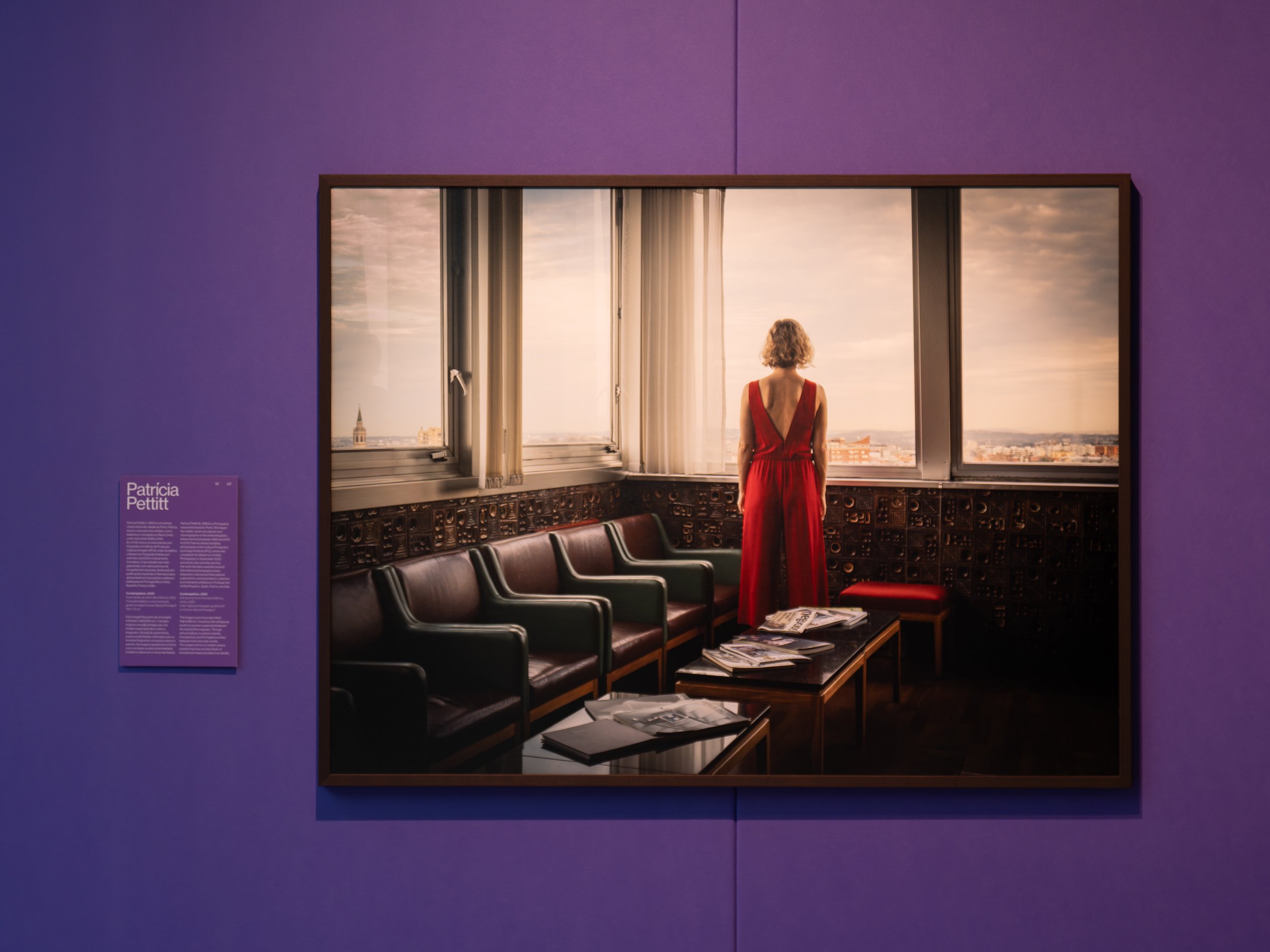 Uma mulher de vestido vermelho observando uma janela ampla com vista para uma cidade ao entardecer. Interior de um espaço com paredes roxas e cadeiras de couro preto.