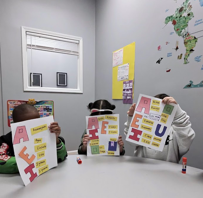 Three children sitting at a table in a classroom holding alphabet posters with words. The classroom has a world map on the wall and a window.