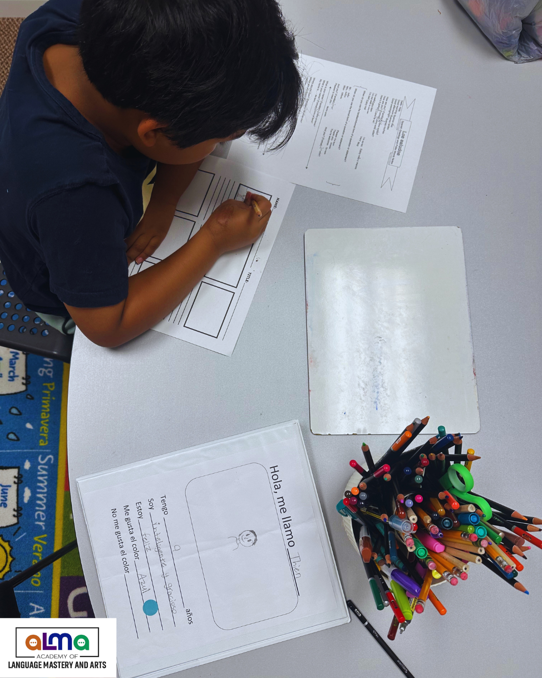 A child working on a language worksheet at a table, with colorful pens in a cup and a whiteboard nearby.
