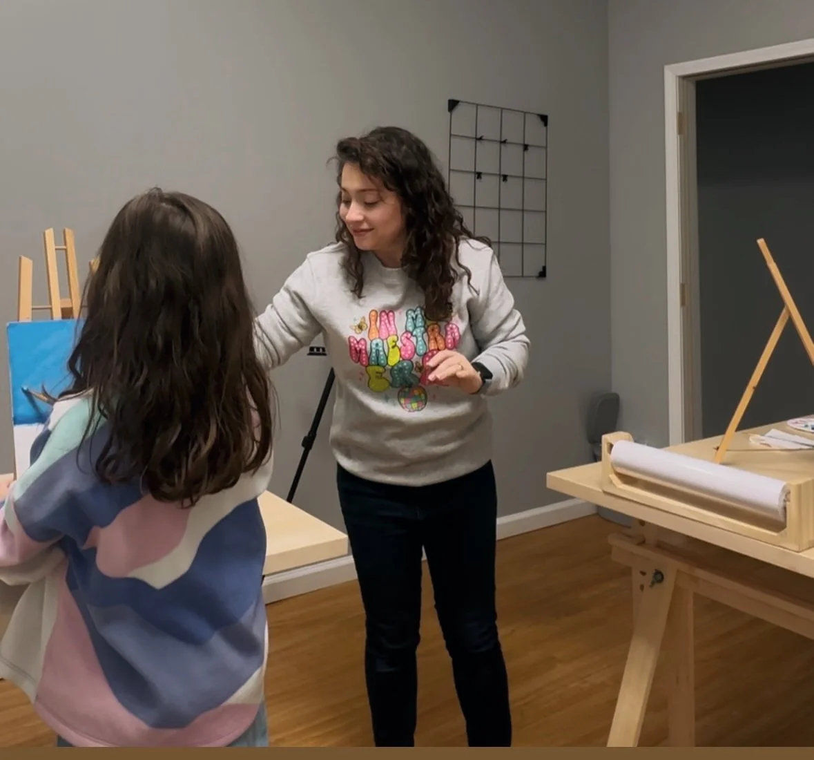 A woman with curly hair wearing a gray sweatshirt with colorful text interacts with a girl with long brown hair in a casual room. There are art supplies and easels.
