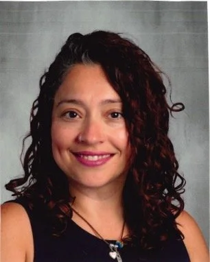 A woman with curly dark hair and a bright smile, wearing a black top and a necklace, standing against a gray background.