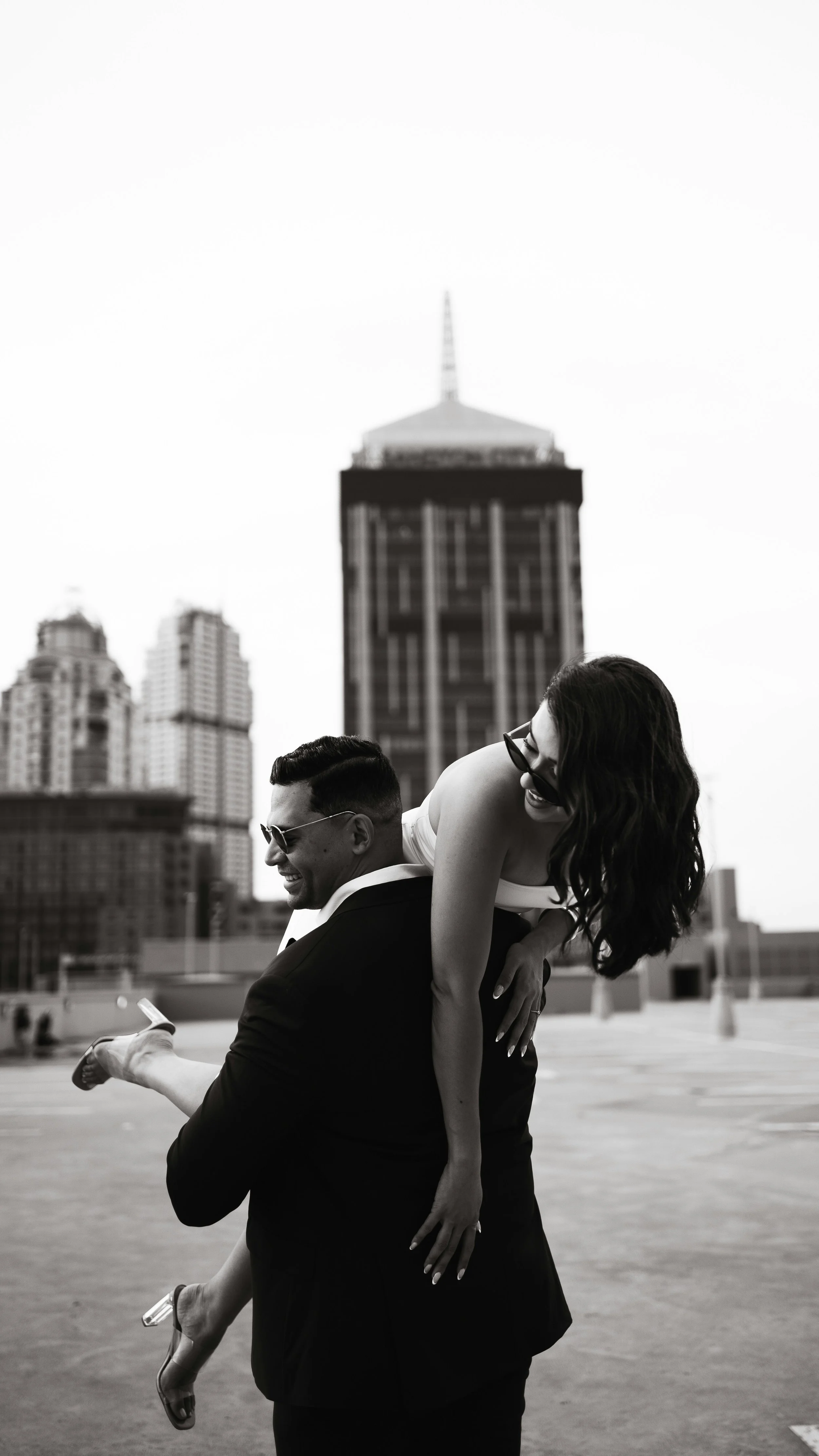 A man in a tuxedo carrying a woman in a white dress and high heels on his shoulders on a rooftop with city skyscrapers in the background.