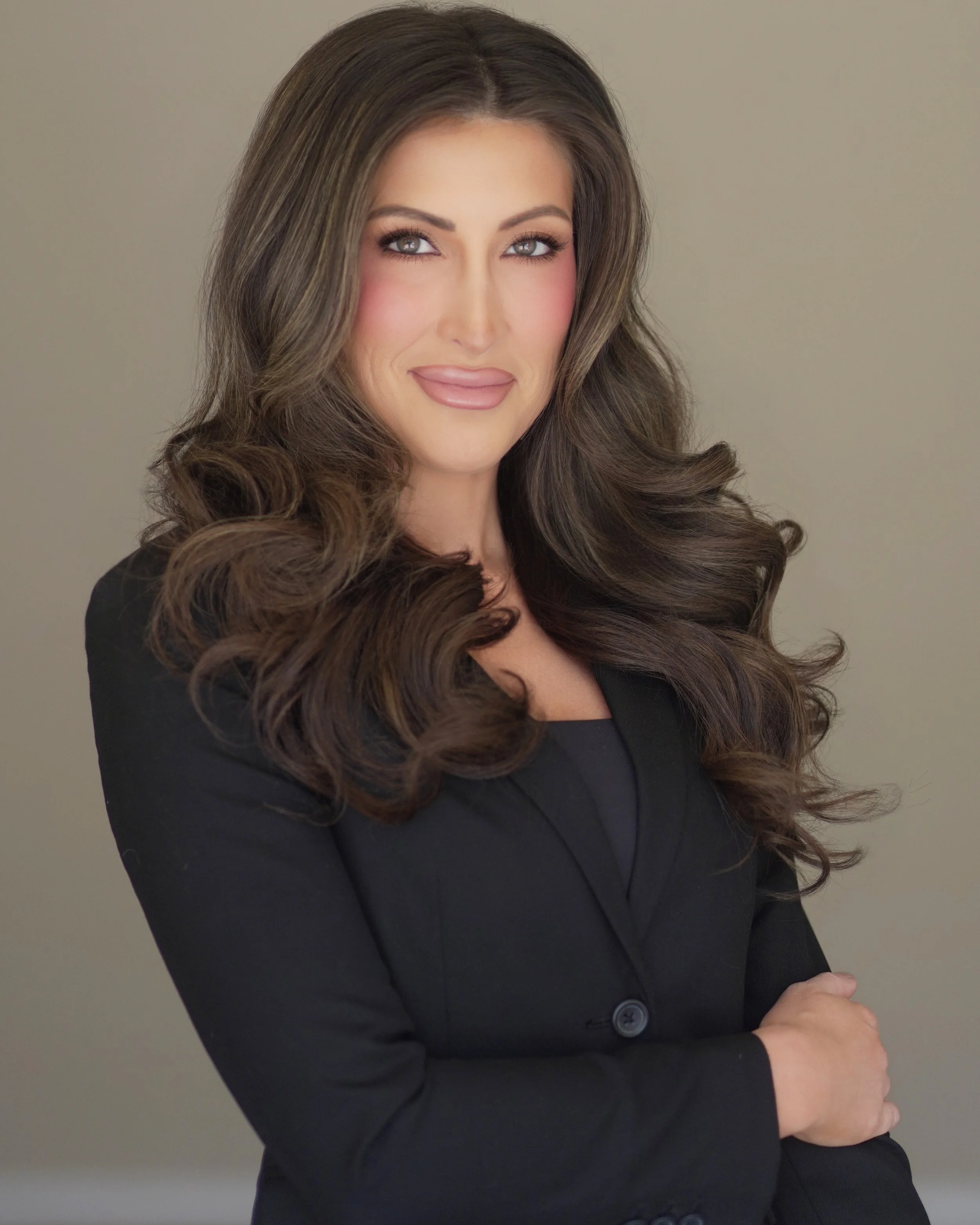 A woman with long, wavy brown hair, wearing a black blazer, smiling with arms crossed against a neutral background.