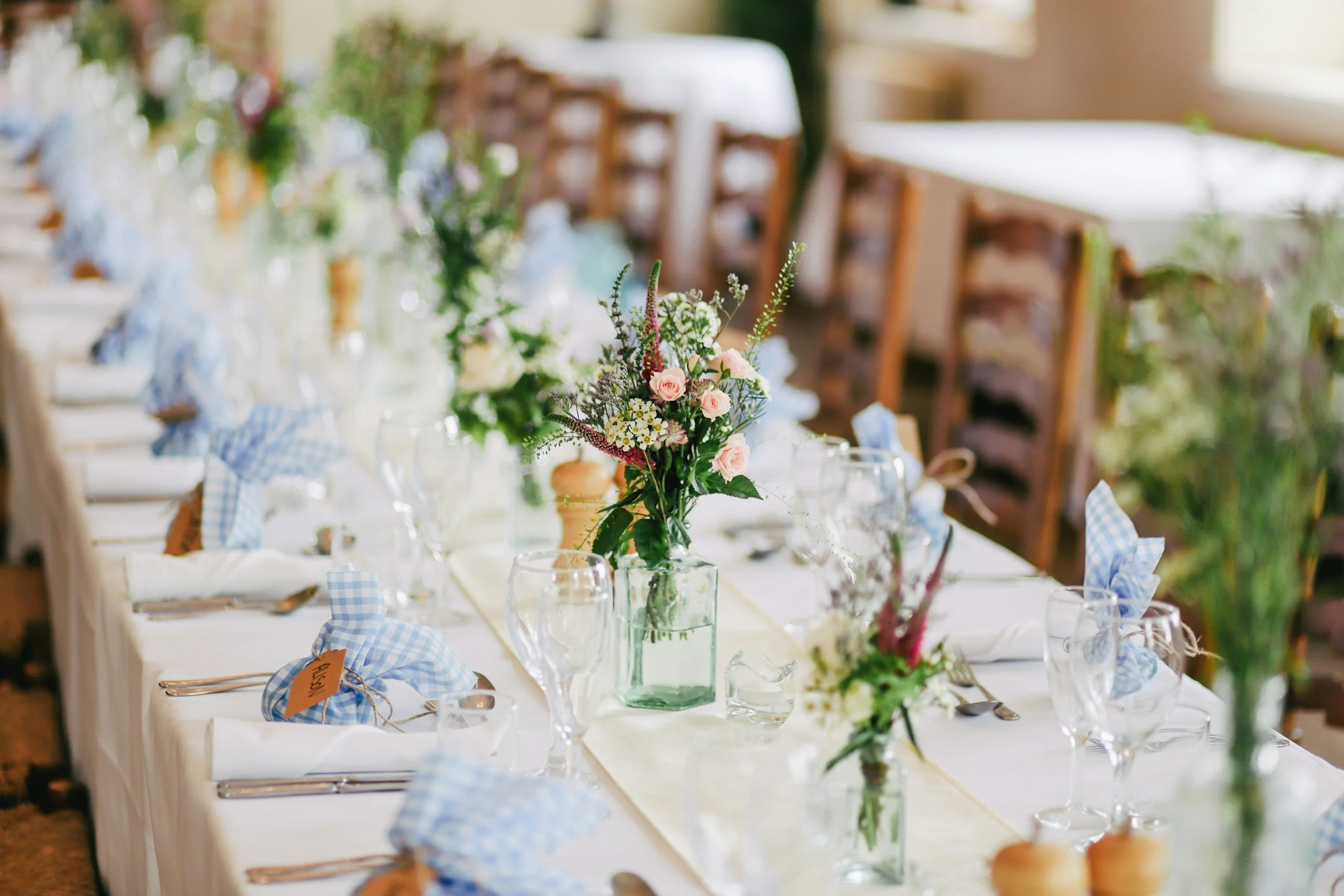 A decorated dining table with a floral centerpiece, blue gingham napkins, wine glasses, and chairs in the background, set in a bright indoor space for a special occasion.
