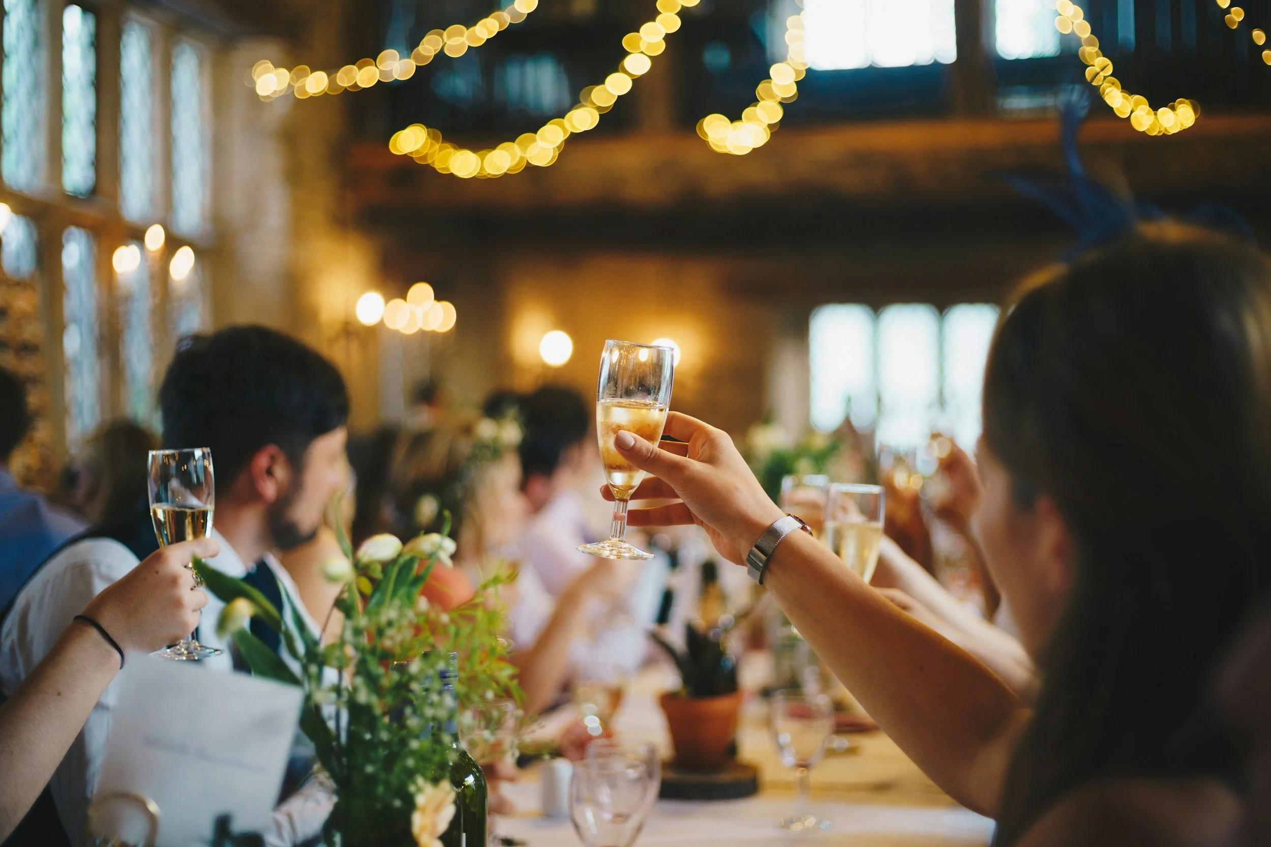 People seated at a dinner table during a celebration or wedding reception, raising glasses of champagne for a toast in a warmly lit rustic venue with string lights.