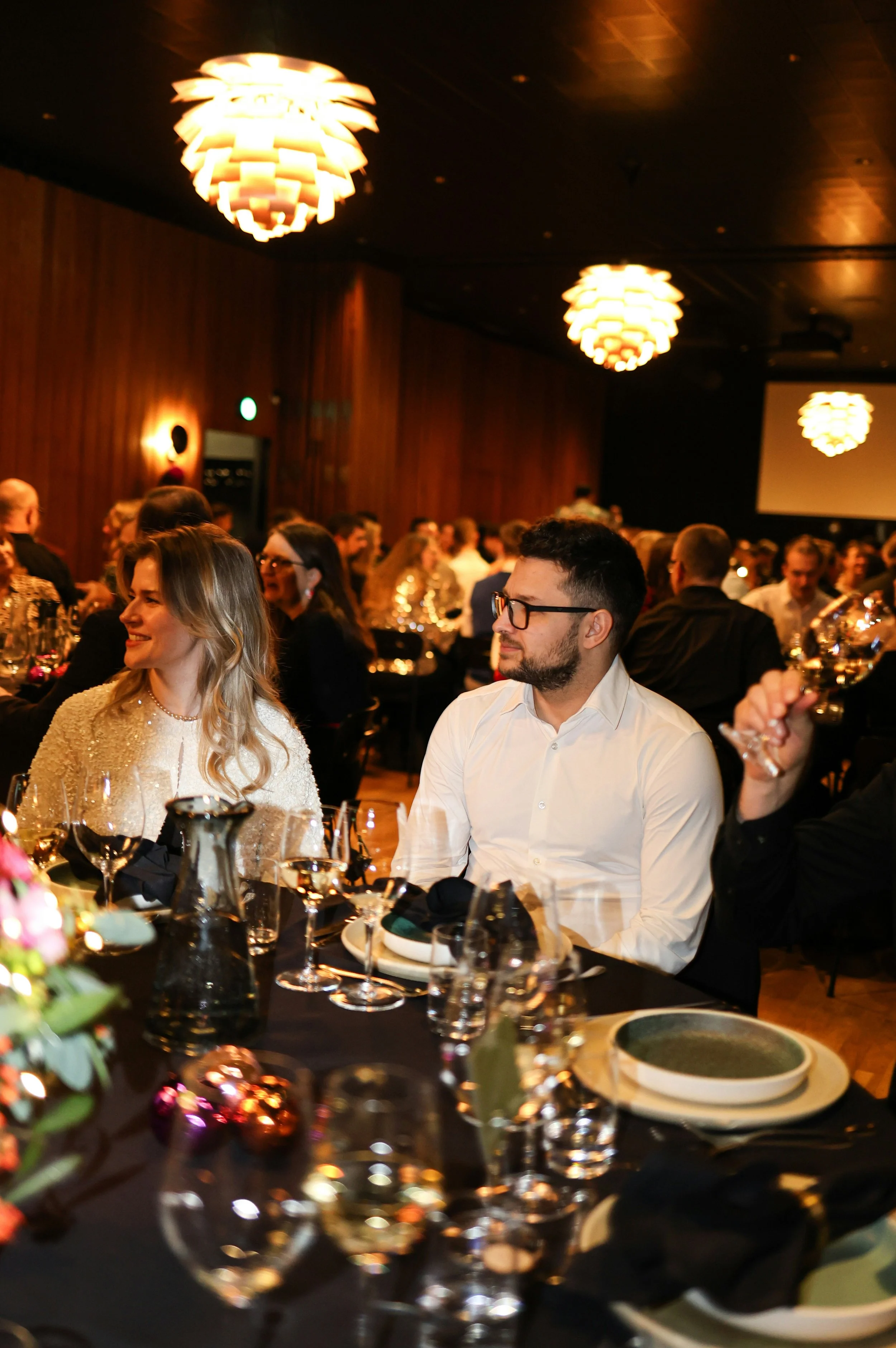 People attending a formal dinner event or celebration in a warmly lit room with wooden walls and decorative ceiling lights.