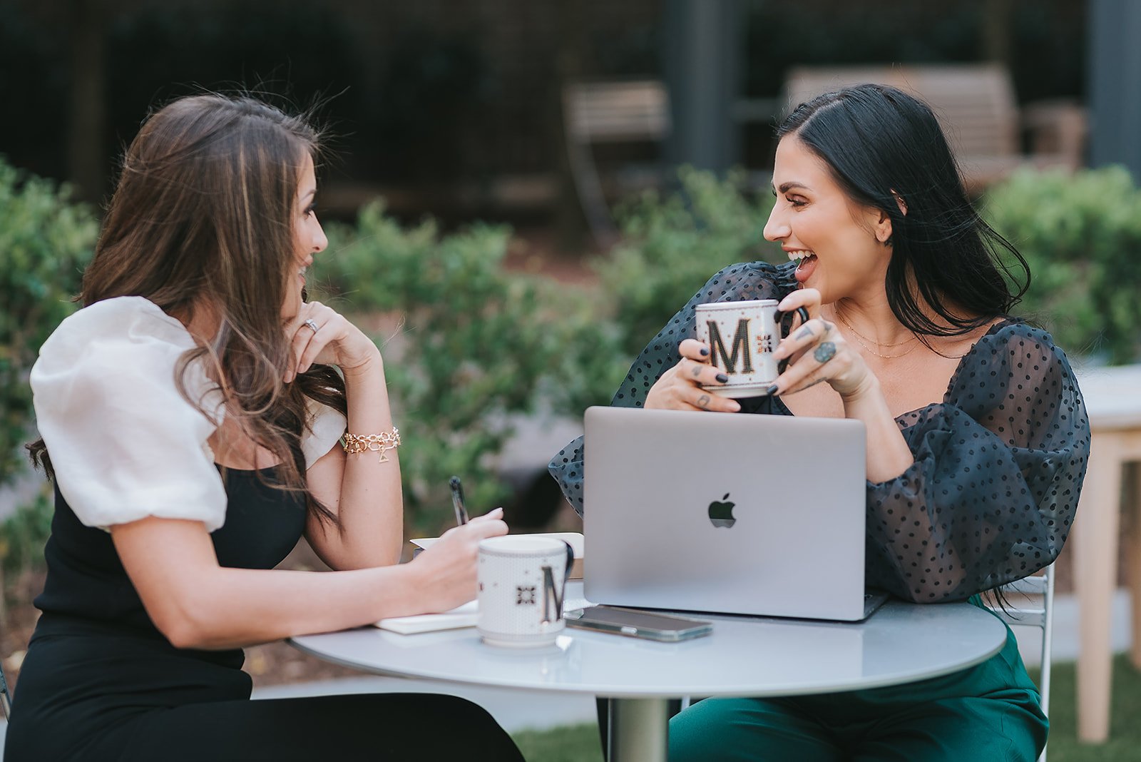 Two women sitting at a outdoor table, having a conversation. One woman is holding a mug and smiling, while the other woman is writing in a notebook. There is a laptop, another mug, and a smartphone on the table.