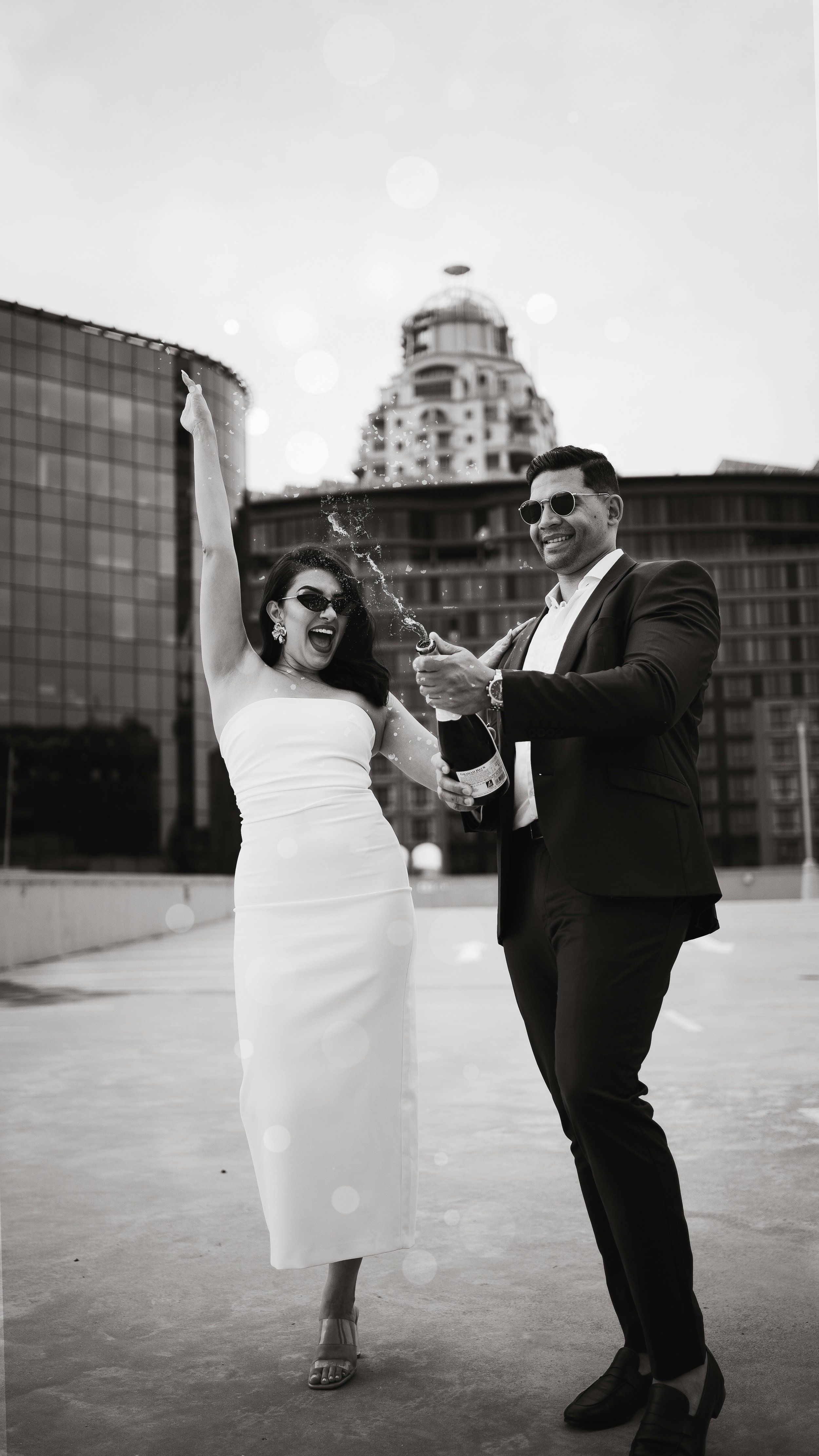 A woman in a white strapless dress and a man in a suit celebrating on a rooftop, opening champagne with skyscrapers in the background.