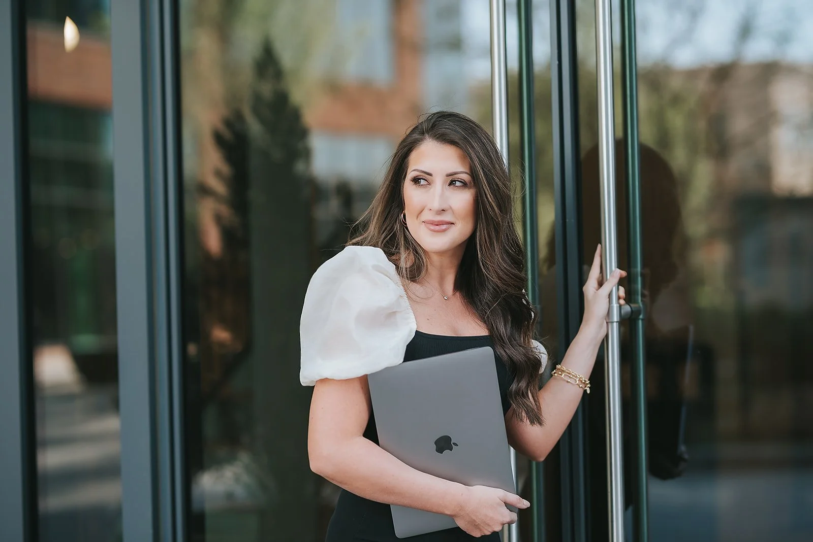 A woman with long dark hair, dressed in a black dress with puffed white sleeves, stands outside near a glass door, holding a laptop and looking to the side.