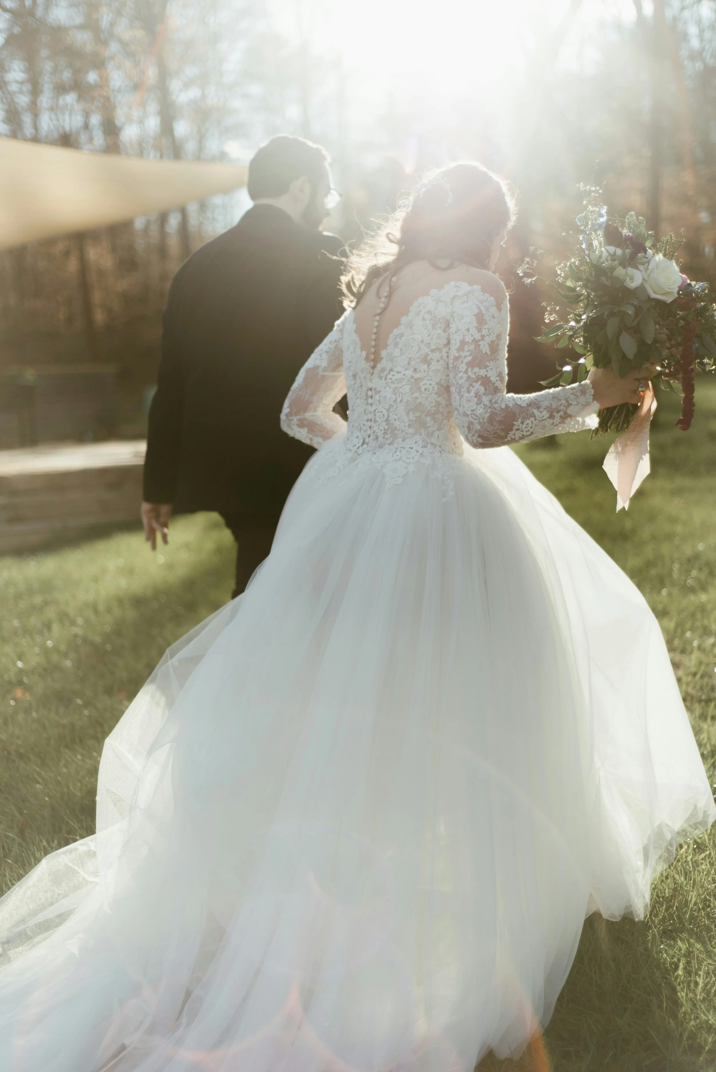 A bride in a lace wedding gown holding a bouquet of flowers, walking outdoors with a groom in a dark suit. The scene is backlit by bright sunlight filtering through trees.