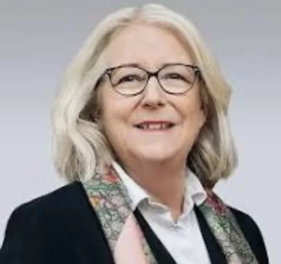 Billedet viser en smilende ældre kvinde med brunt hår og briller, iført en sort jakke og en farverig halsedisse.
