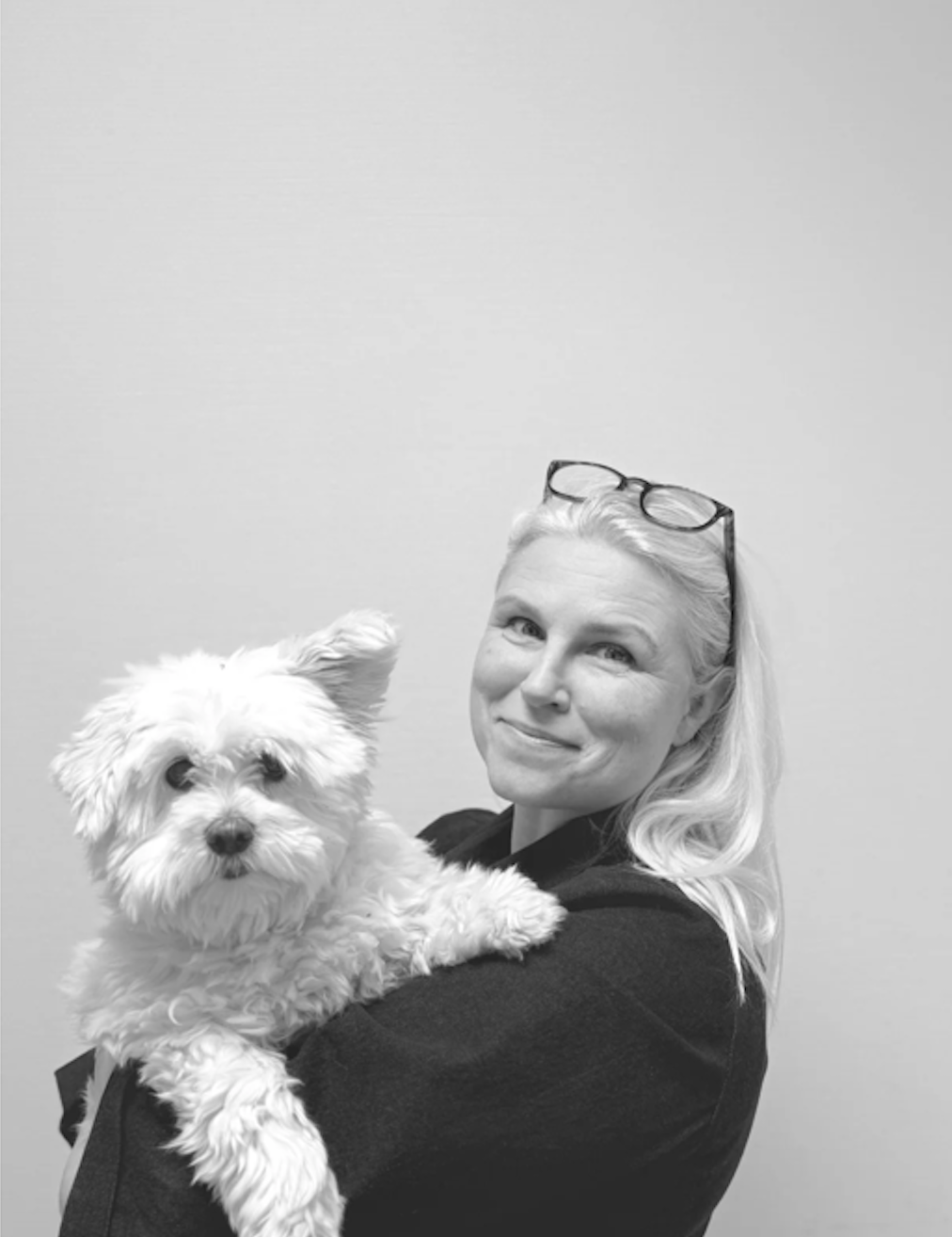 En kvinde holder en lille, fluffy, hvid hund i sine arme, de står foran en neutral væg, sort-hvid foto.