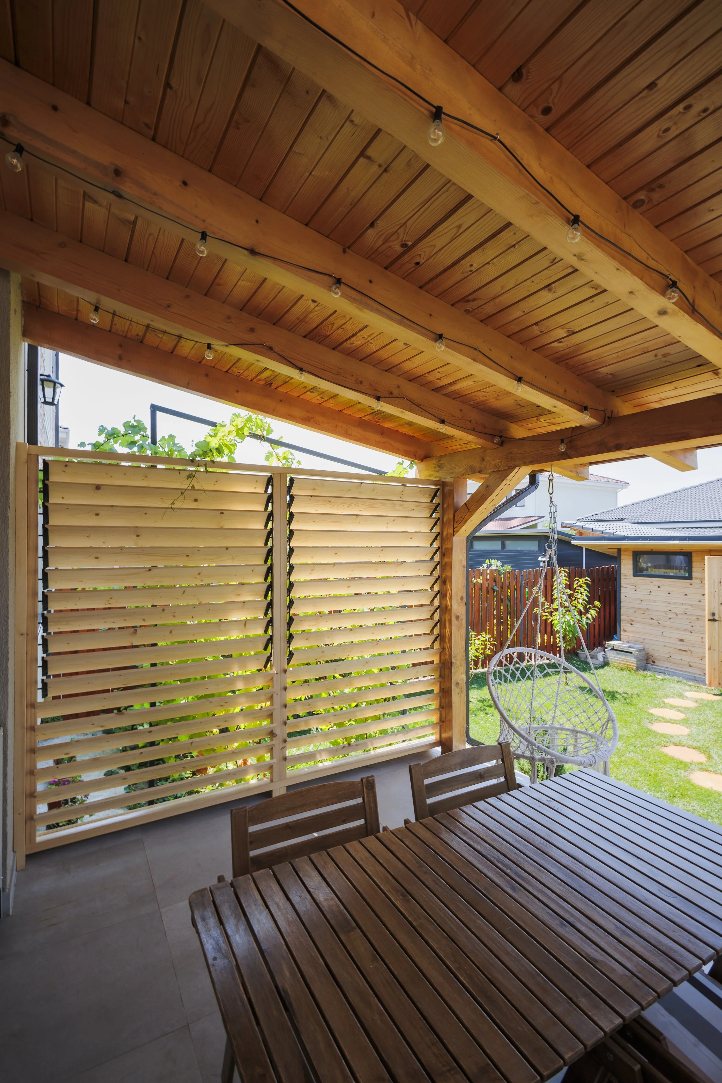 A wooden patio cover with string lights, a slatted privacy screen, outdoor furniture including a wooden table and chairs, a hanging egg chair, and a view of a backyard with a hammock and garden.