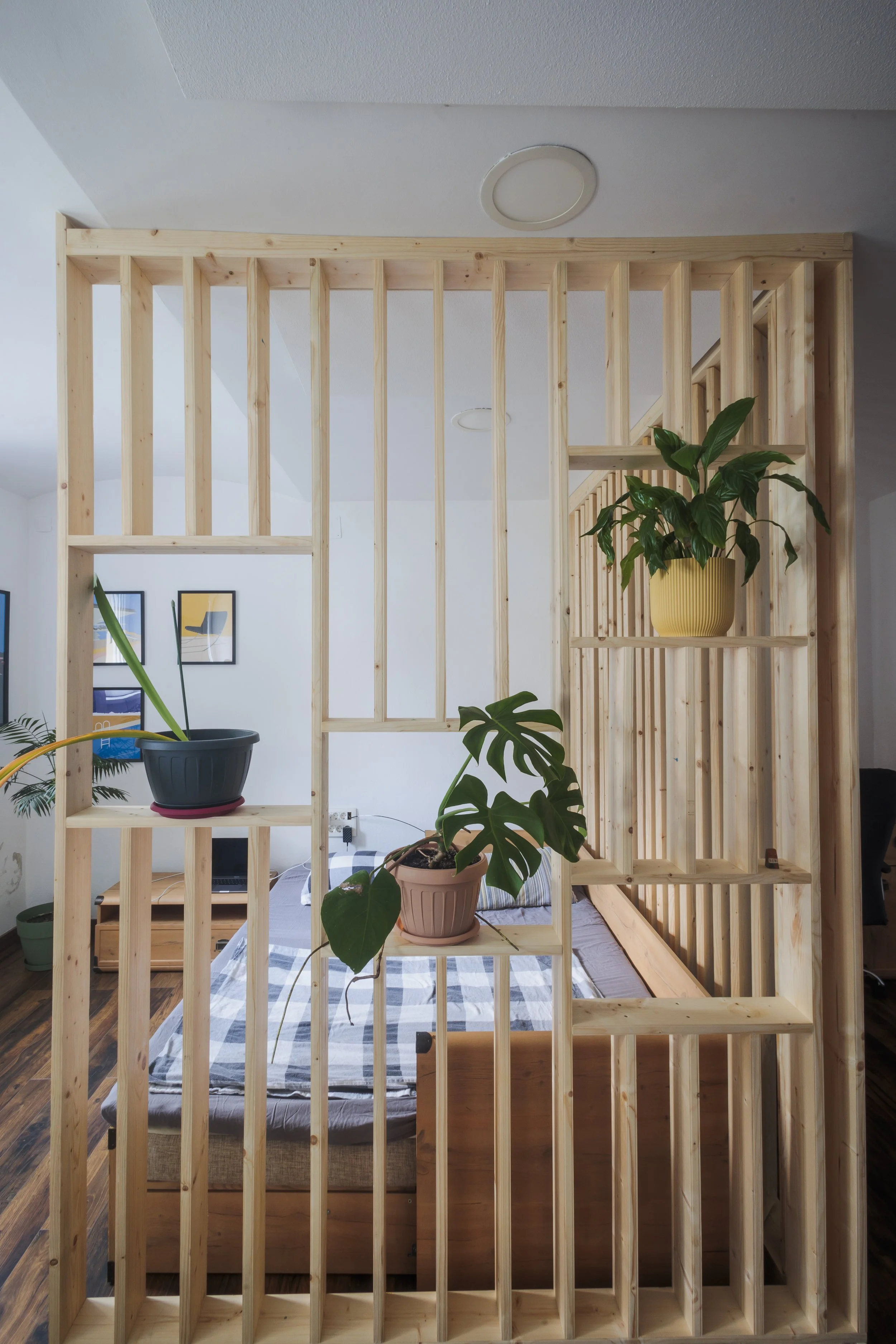 Interior view of a wooden room divider with vertical slats, separating a sleeping area with a bed, potted plants, and framed pictures on the wall.