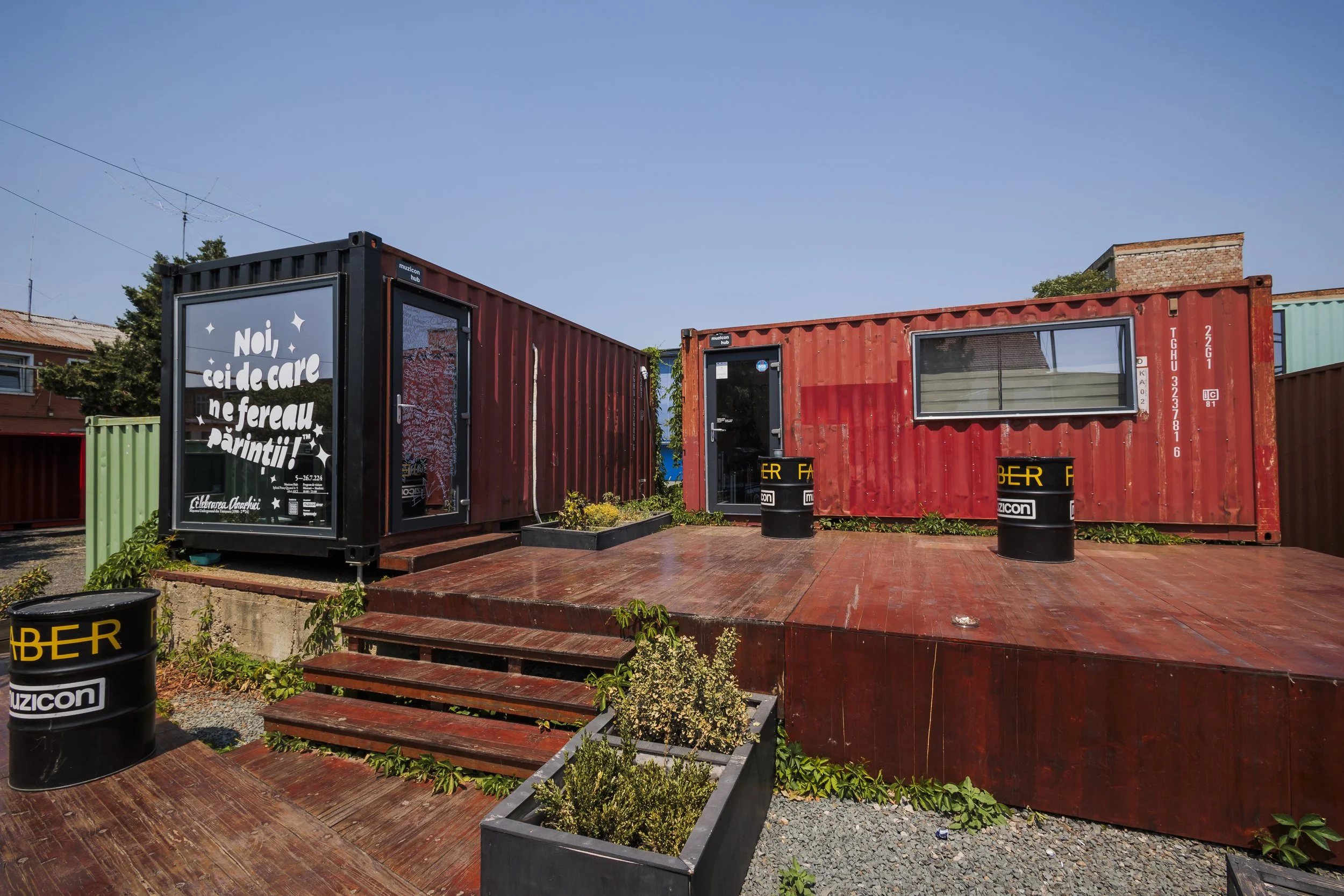 Outdoor scene with the sky visible and old shipping containers repurposed as buildings or shops, some with large windows and doors, surrounded by planters and barrels, with a wooden deck and steps leading up to it.