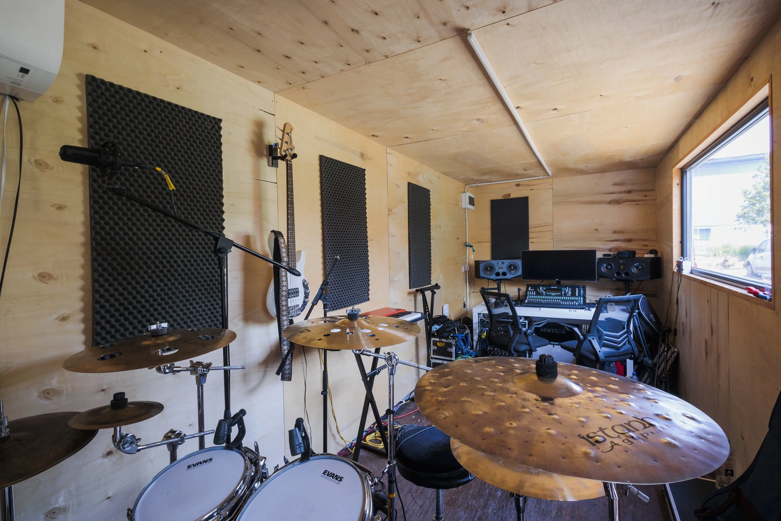 A music studio with wooden walls and ceiling, containing a drum set, guitars mounted on the wall, a keyboard, computer monitors, speakers, and soundproof foam panels.