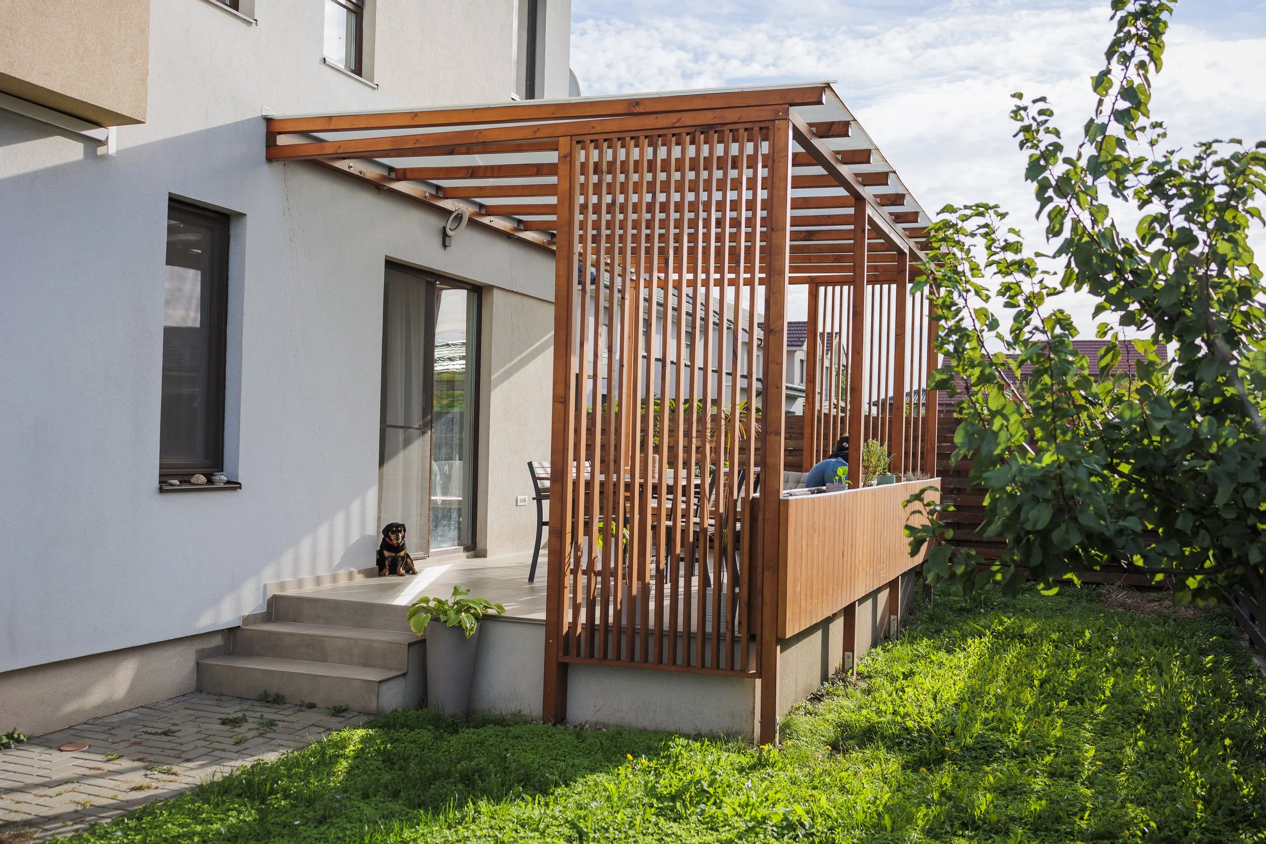 A backyard patio with a white house wall, a small black and brown dog sitting by the steps, and a wooden pergola structure with slatted roof and vertical side panels. There are potted plants and outdoor furniture on the patio, and a grassy lawn with trees in the background.