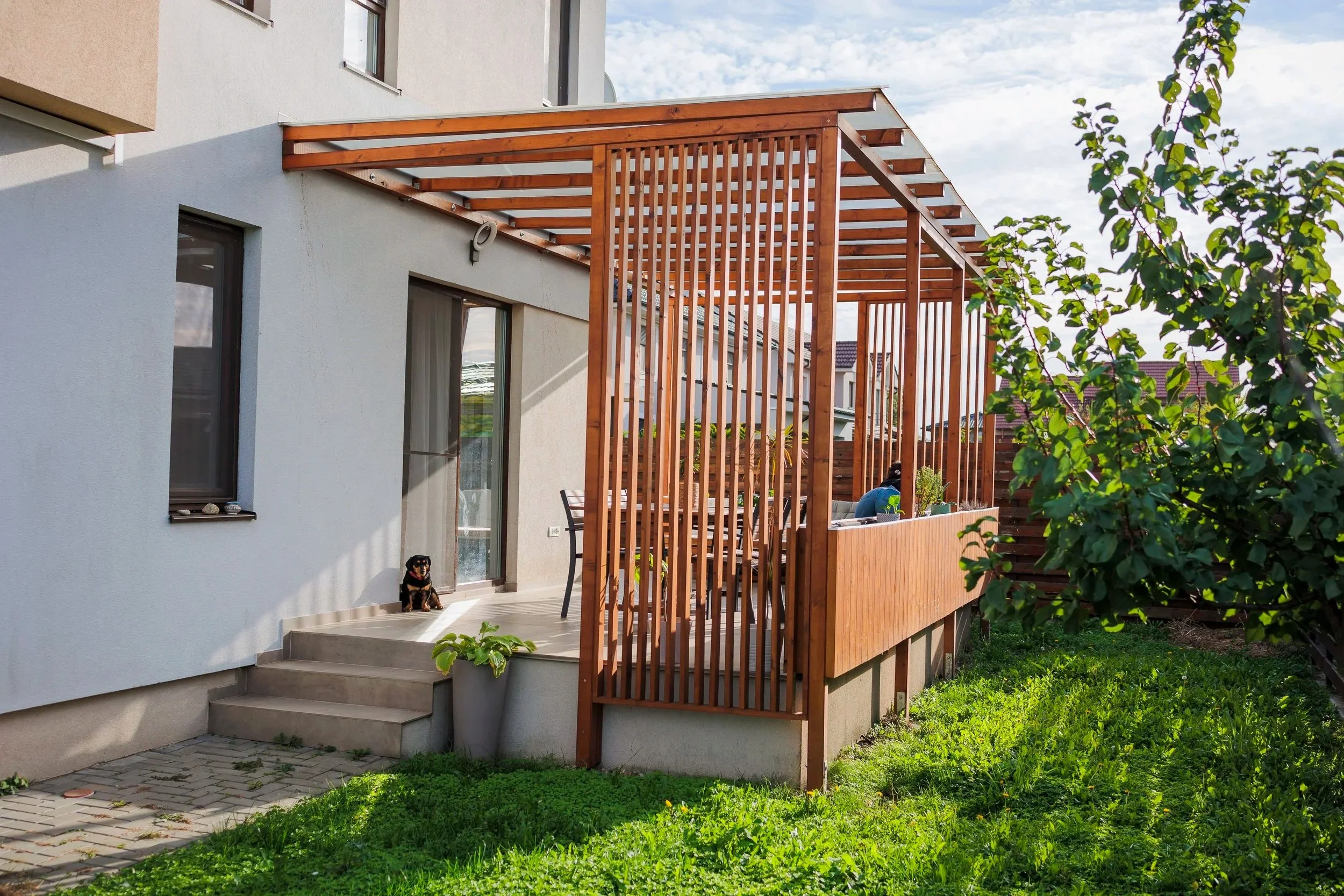 A backyard patio area with a white building, sliding glass door, gray steps, and a black and tan dog sitting on the steps. There is a wooden privacy fence and a partially constructed wooden pergola or deck extension. Green grass and trees are visible