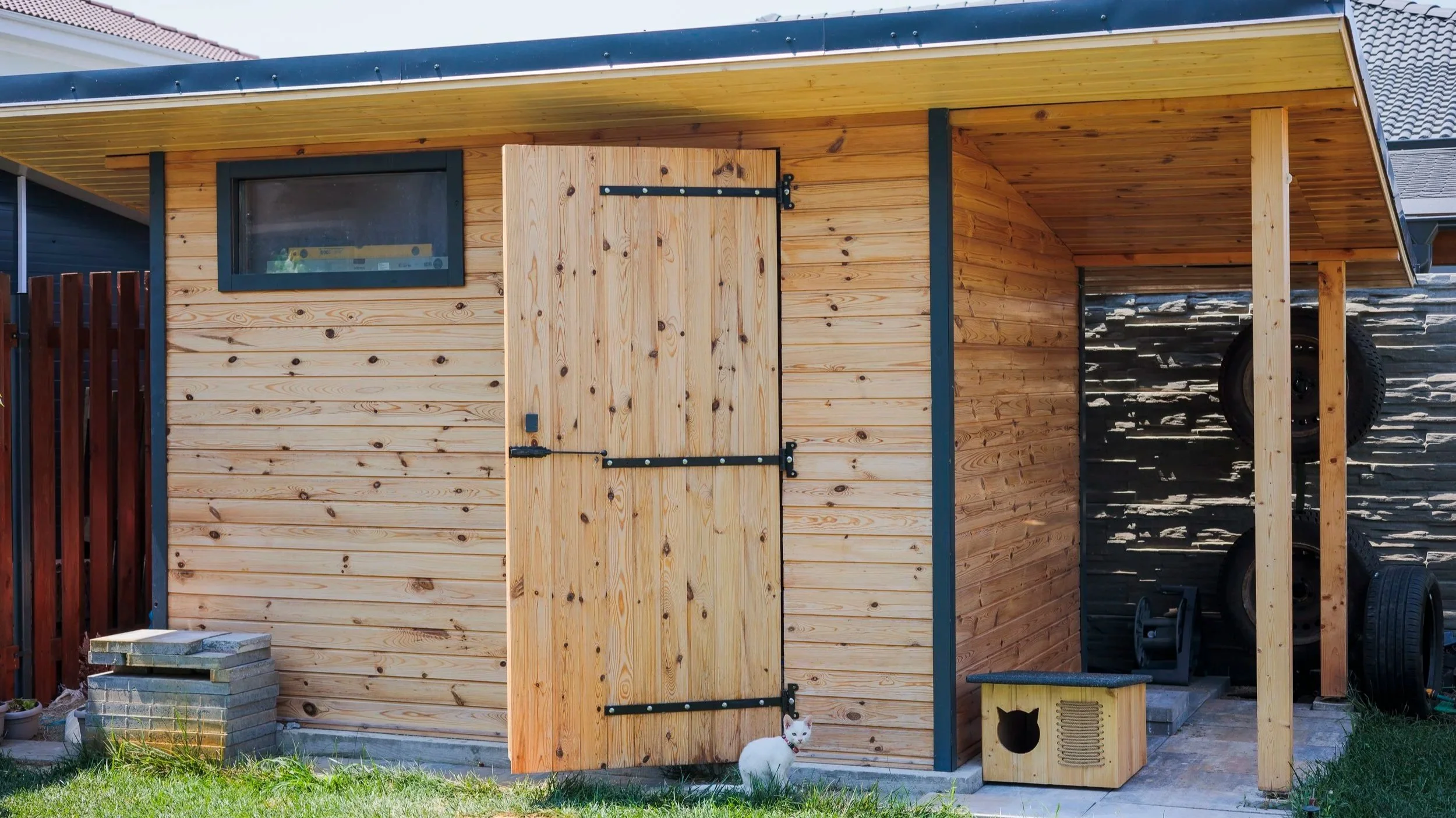 Wooden outdoor structure with a door, window, and a small animal shelter, situated on grass with stacked bricks nearby.