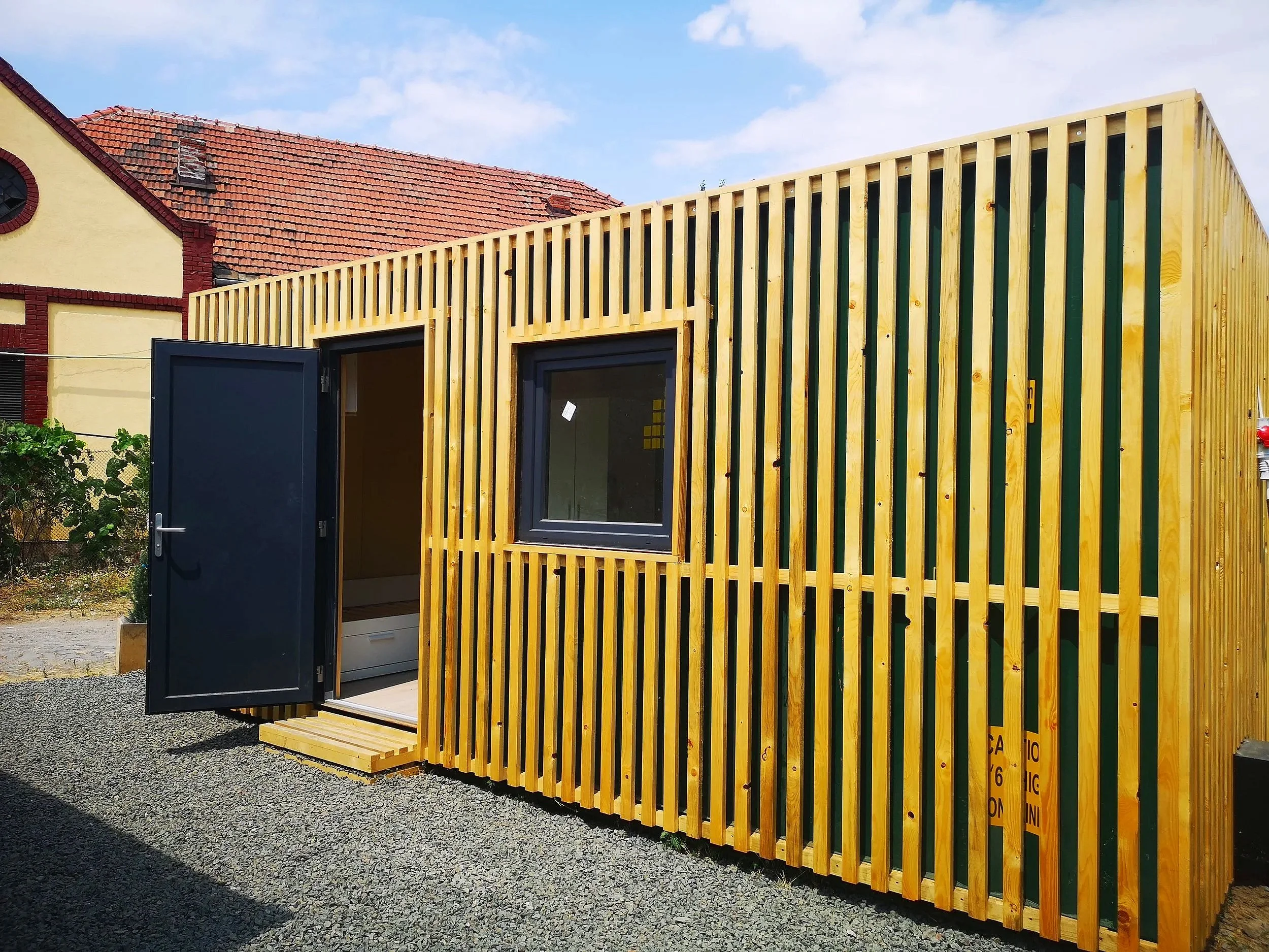 A small wooden structure with vertical slats and a black door, attached to a red-tiled building with a small window in the roof, set against a blue sky with clouds.