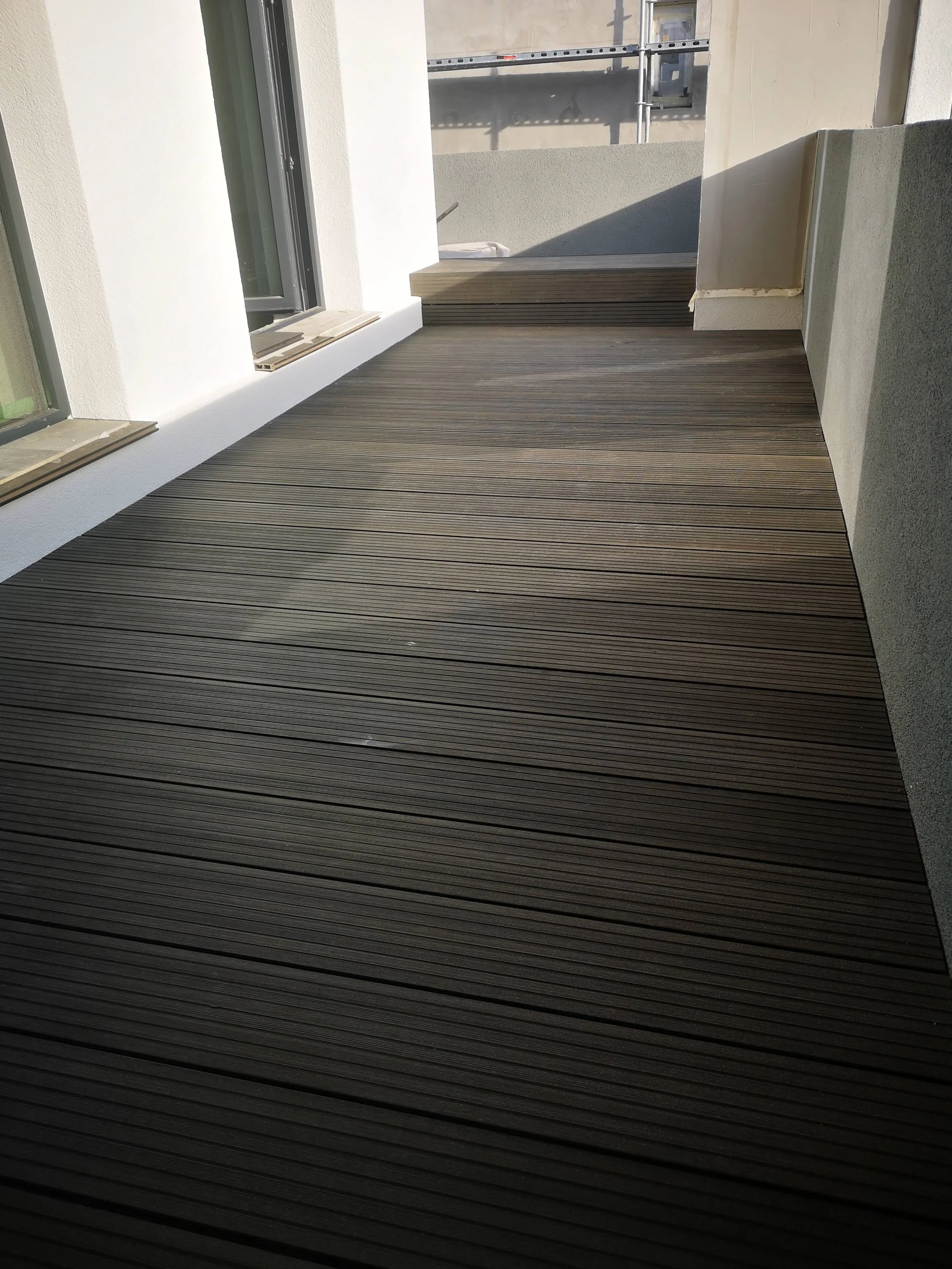 A modern outdoor balcony or terrace with wooden deck floor, white walls, and a large window. There is a small ledge or step at the end of the balcony, with some construction scaffolding visible in the background.