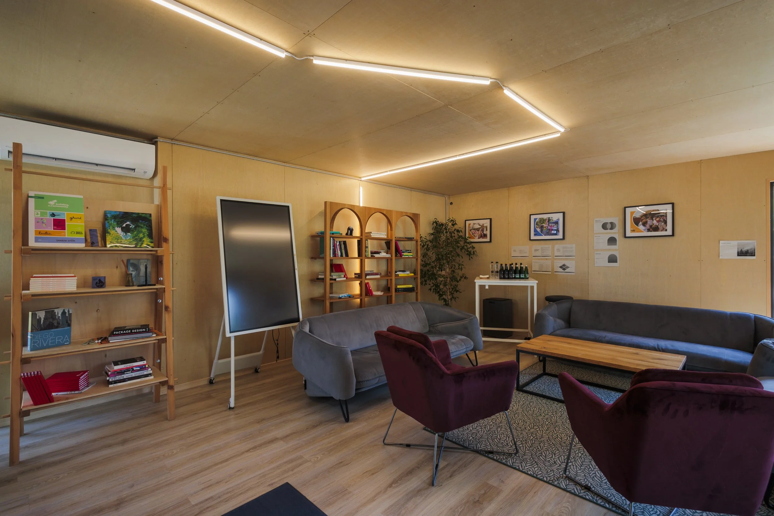 A cozy, modern lounge area with several comfy chairs and a wooden coffee table. The walls are decorated with framed pictures and diagrams, and a bookshelf with books and colorful artwork stands against the wall. The ceiling features unique lighting f
