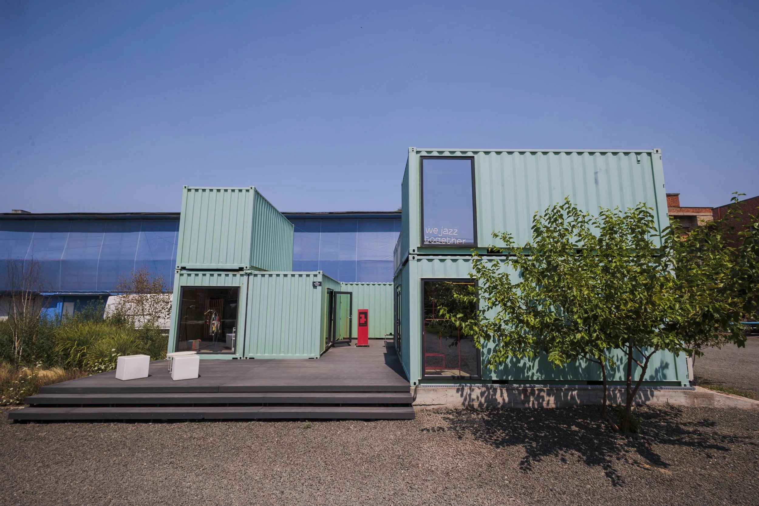 A modern building made of stacked shipping containers painted in light green, with open doors and windows, surrounded by trees and greenery, under a clear blue sky.