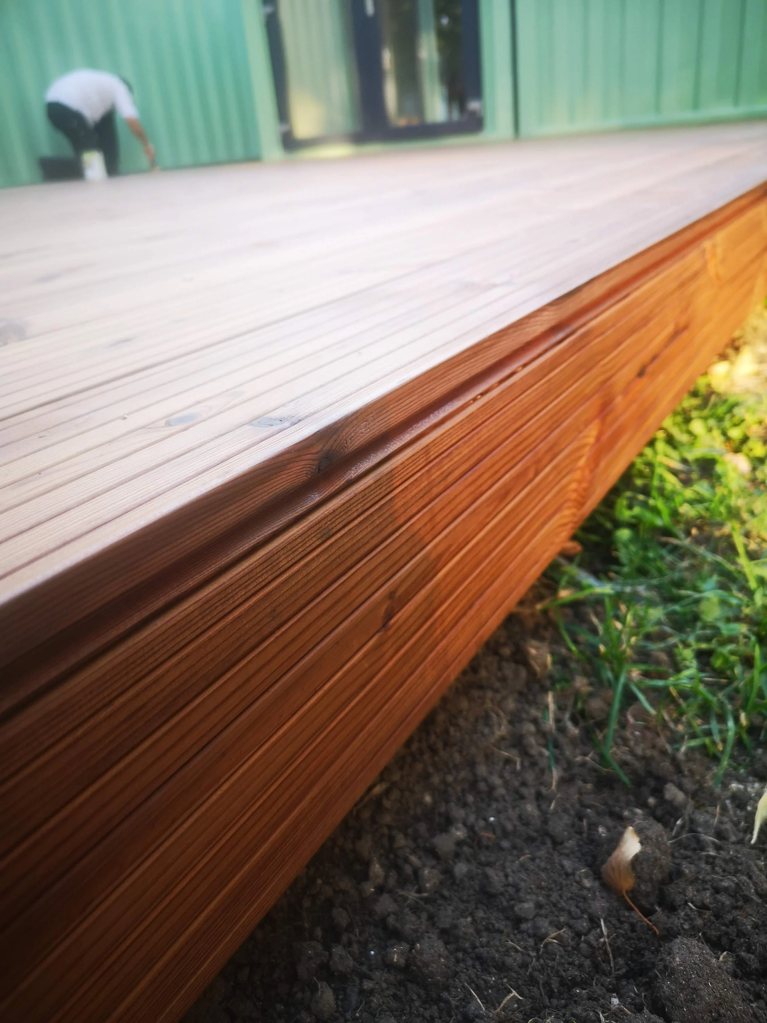 Close-up of a wooden deck edge with grass and soil underneath, and a person painting a green building in the background.