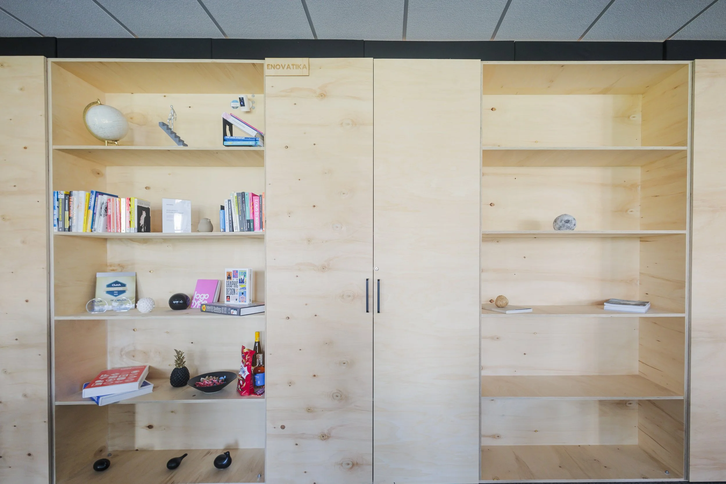 A large wooden shelving unit with five open shelves and a closed cabinet at the bottom. Items on the shelves include books, decorative objects, a globe, a small sculpture, and various ornaments. The setting appears to be an office or study space.