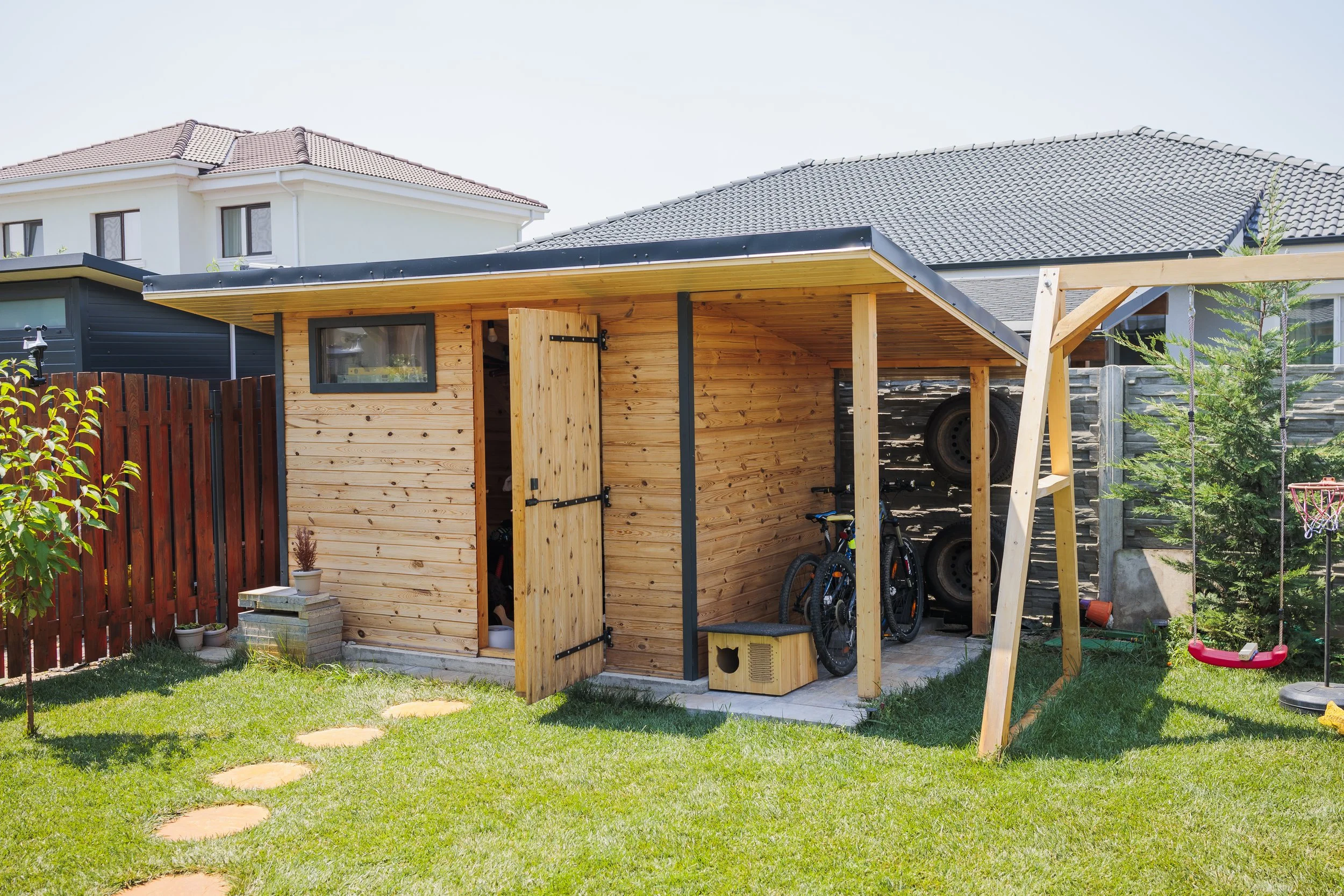 Backyard with small wooden shed, garden beds, bicycles, and a play swing set, with neighboring houses visible.