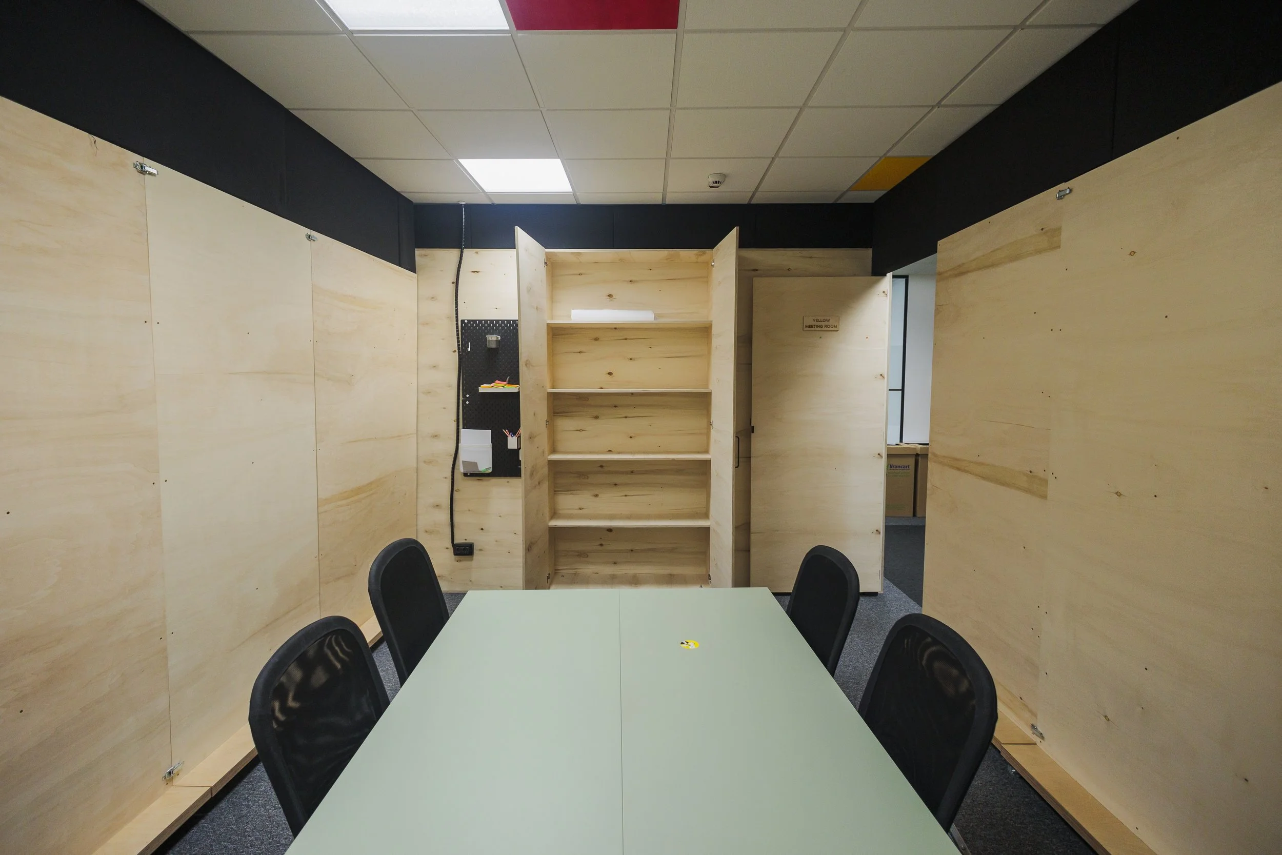 An office space with a green table surrounded by four black chairs. The walls are made of unfinished wood panels, and the ceiling has white and red tiles. A small bookshelf is visible behind the table, and there is a door with a sign that reads 'Yell