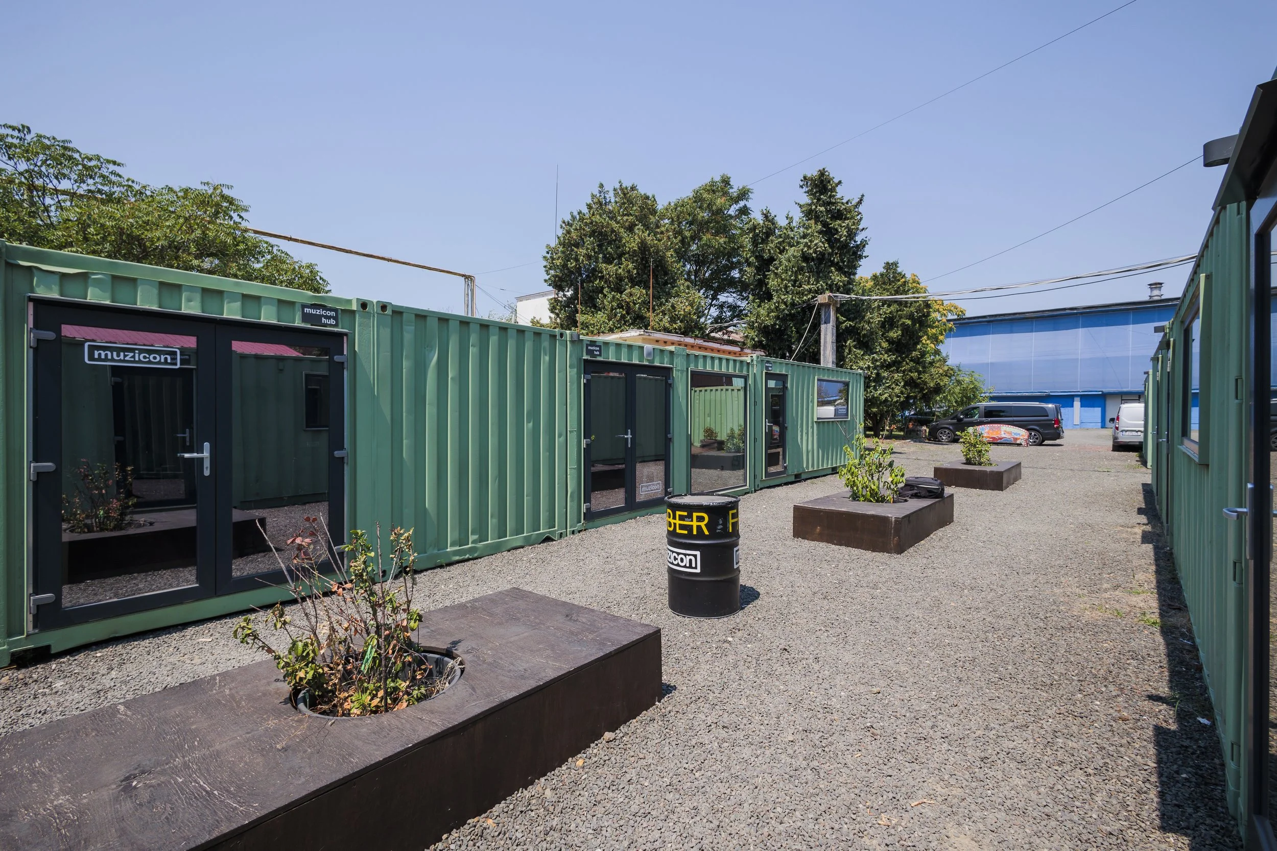 Outdoor scene with green converted shipping containers used as storefronts or offices, some with signage reading "muzicon." There are trees, a parking lot with cars, and a blue building in the background, under a clear blue sky.