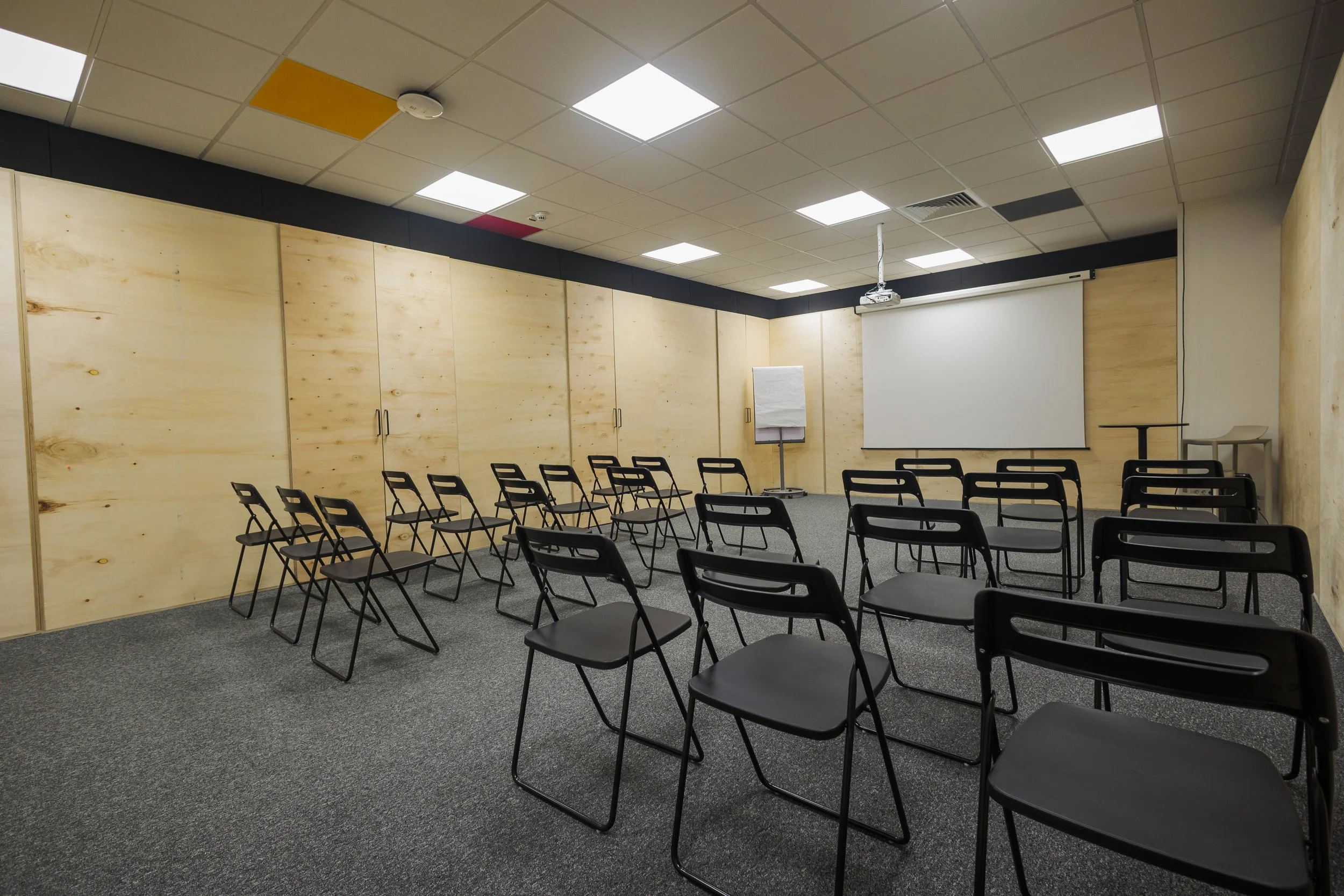 Empty conference room with rows of black chairs, a white projection screen, and a flip chart with paper on an easel. Wooden wall panels and ceiling lights are visible.