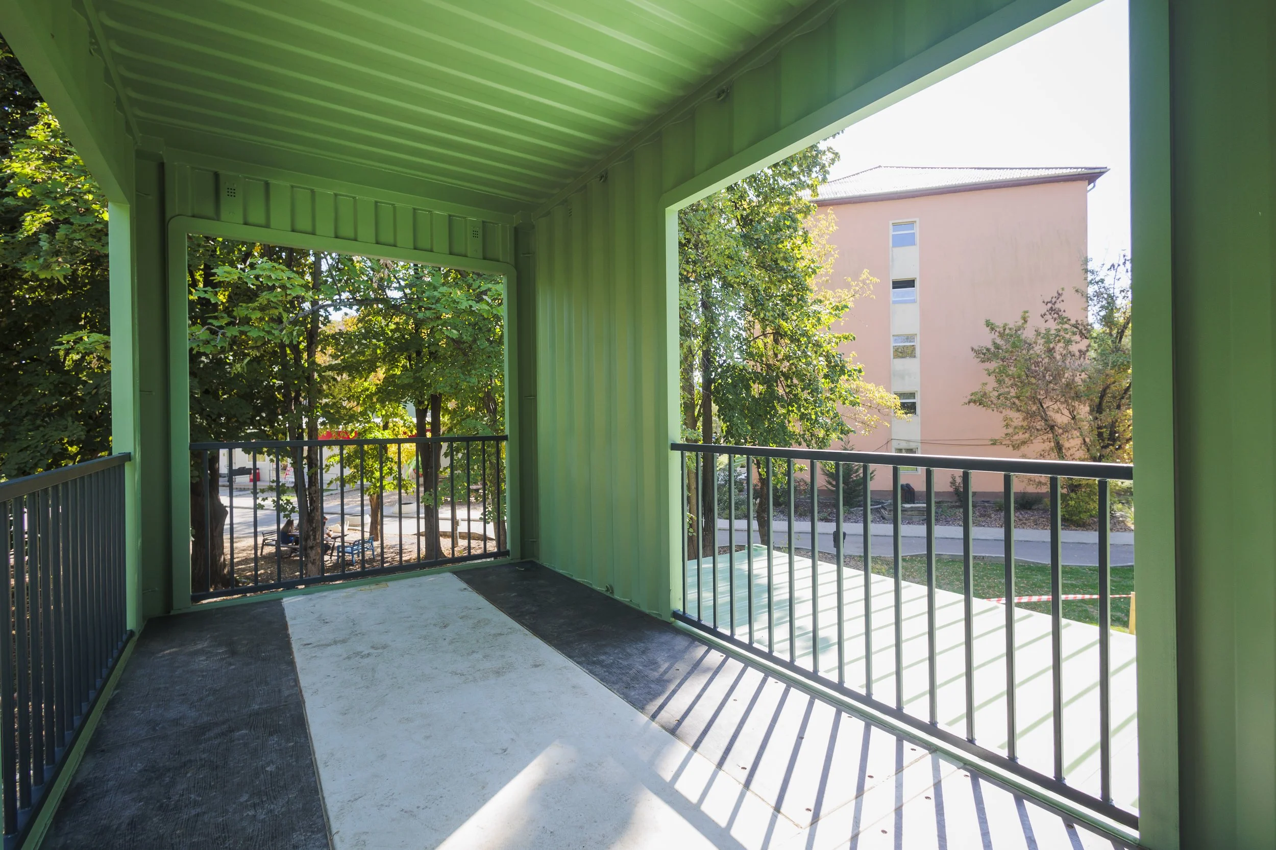 View from a balcony with green-painted metal walls, railing, and a ceiling, overlooking a tree-lined street and a peach-colored building.