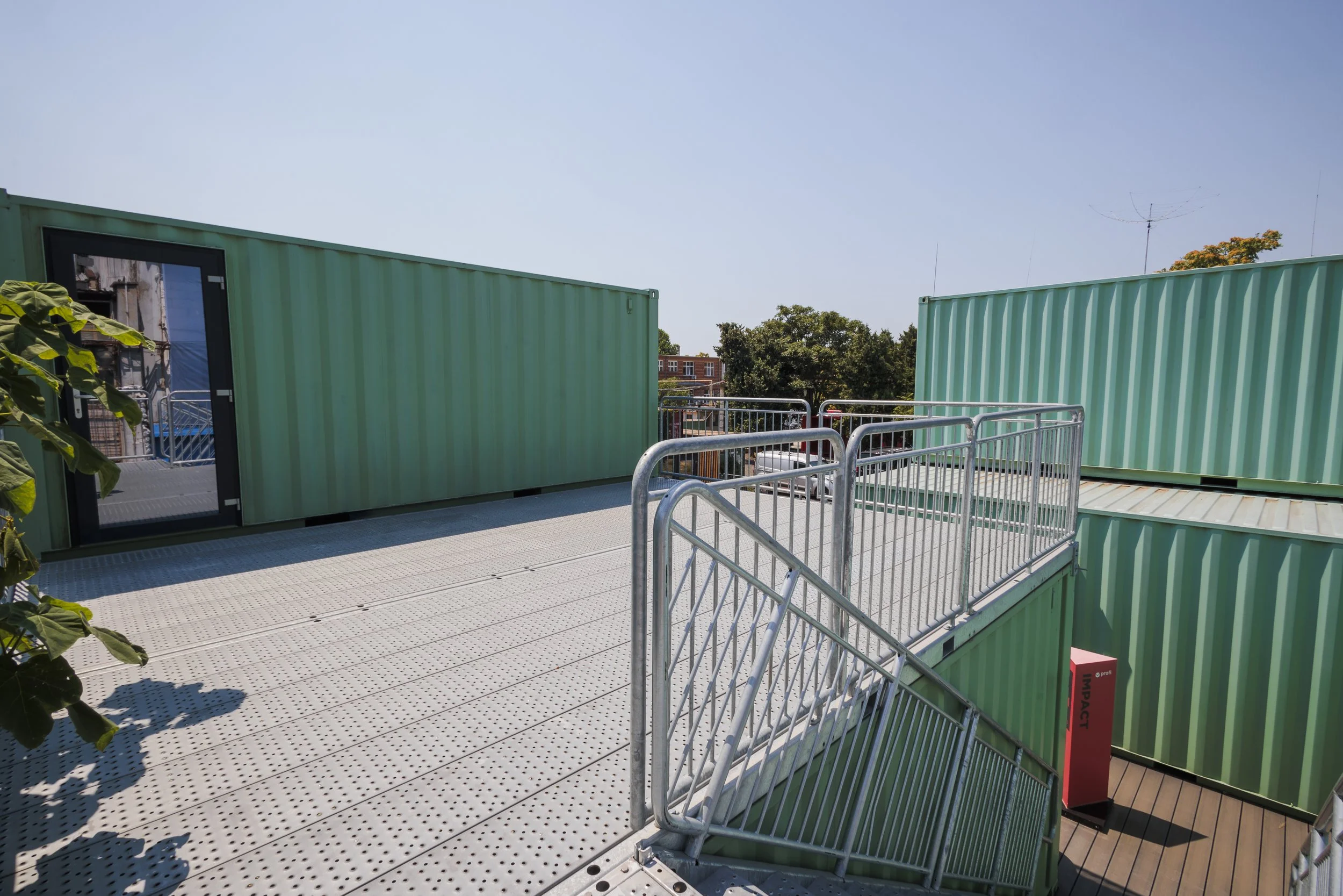 Outdoor rooftop with metal railings, large green shipping containers, and a door leading inside. Clear blue sky with trees and buildings in the background.