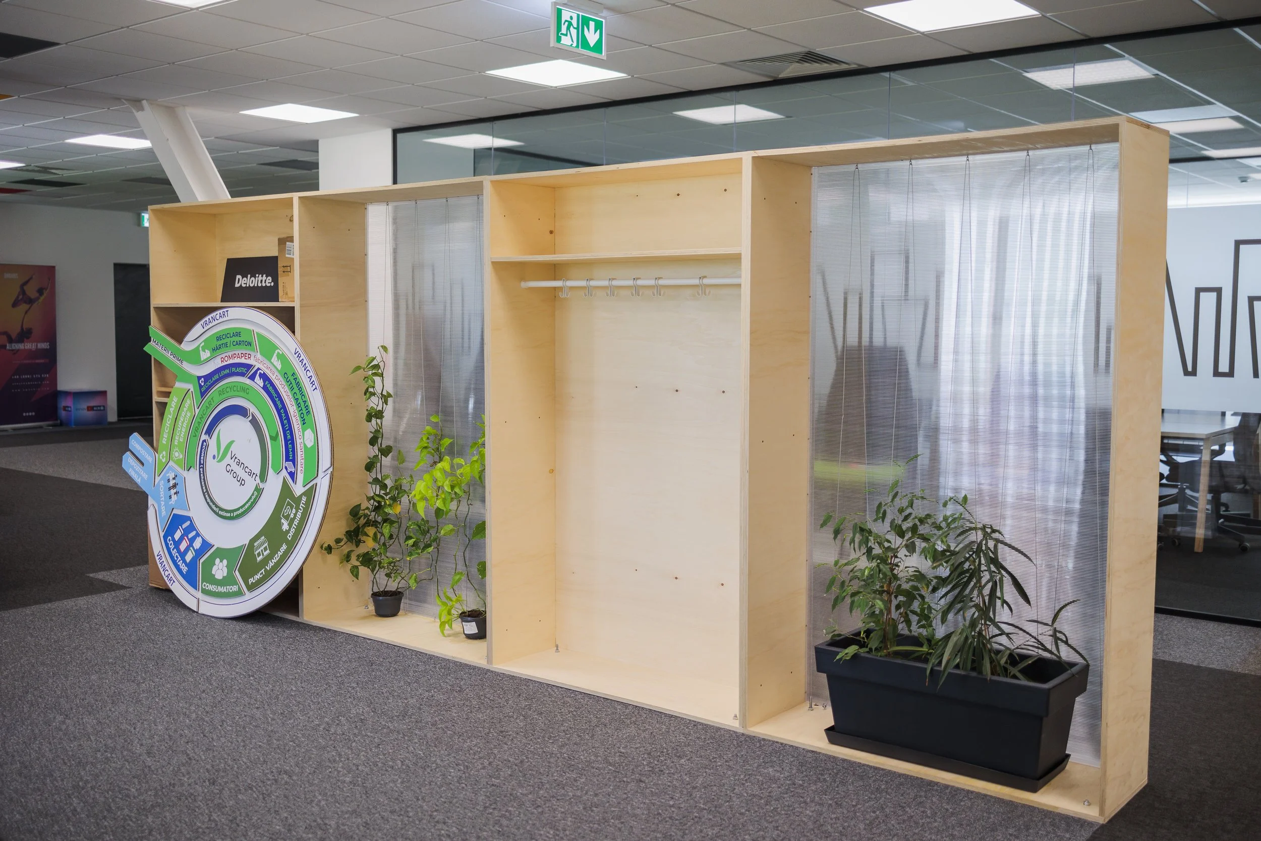 Empty wooden display booth with transparent plastic curtains, green plants, and a circular informational sign in an office or store environment.
