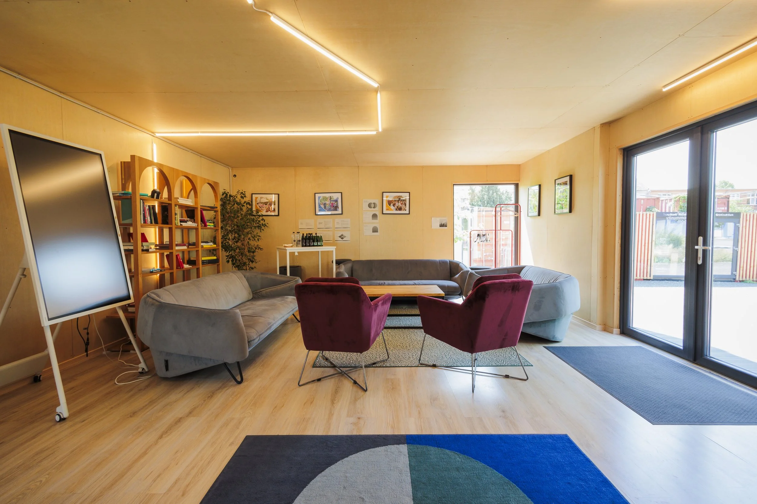 A modern lounge area with wooden floors, beige walls, large windows, and ceiling lights. There are several sofas, a bookshelf, framed artworks, a large mirror, and a colorful rug.