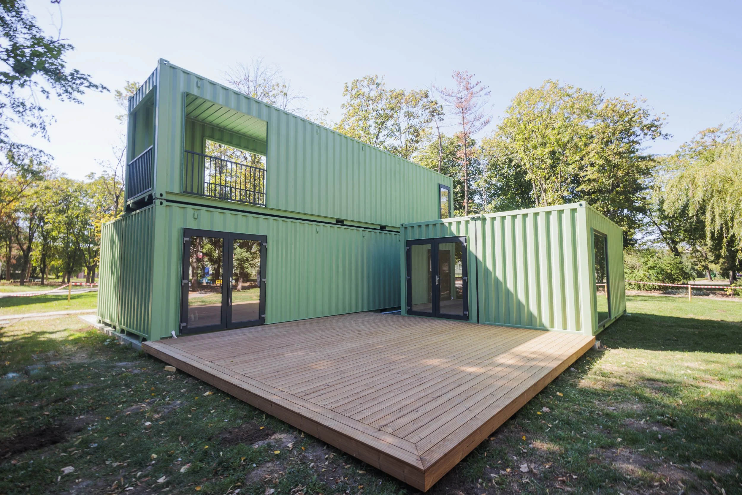 Green container home with large sliding glass doors and a second-floor balcony, set on a wooden deck in a park surrounded by trees.