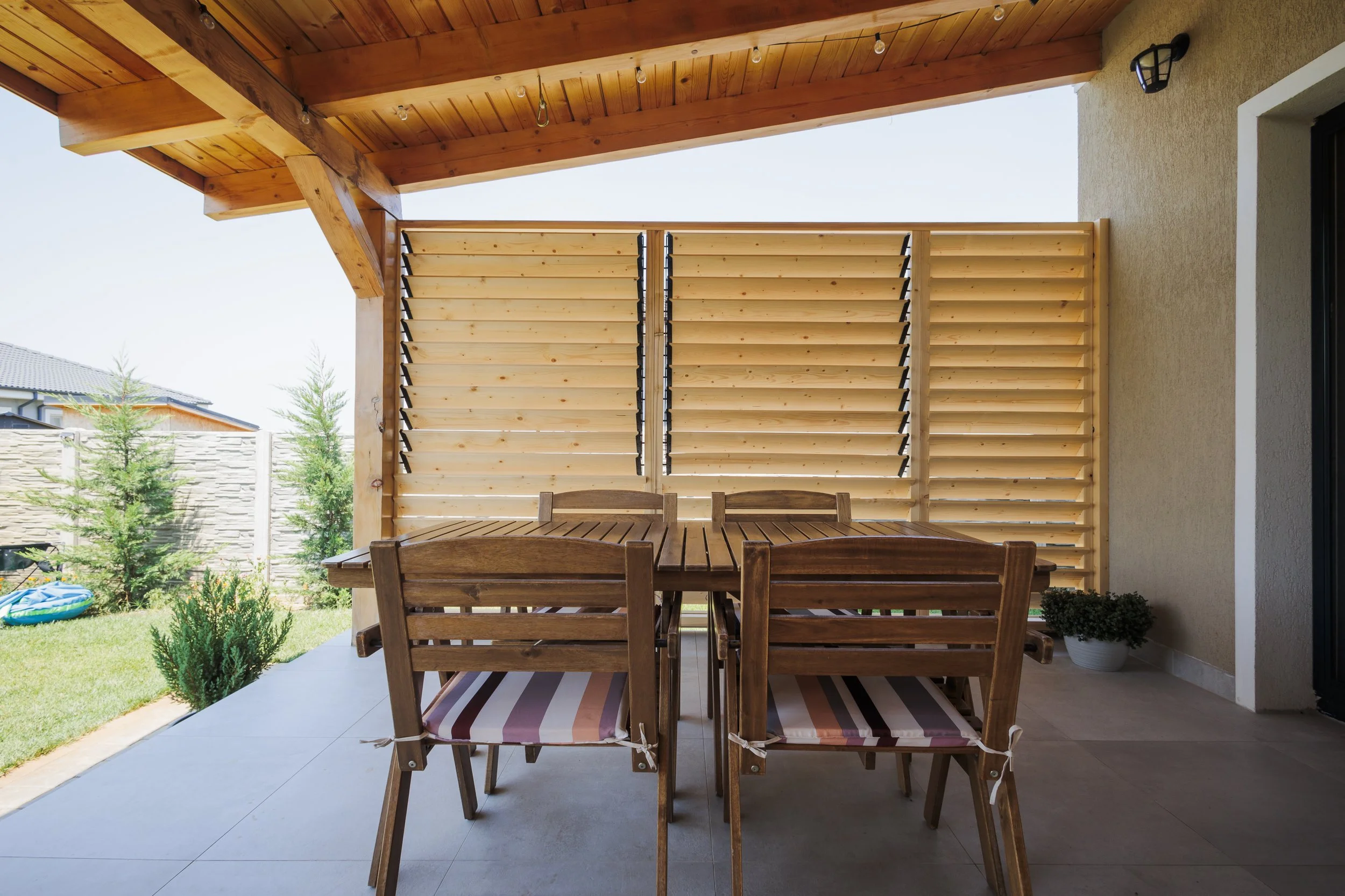 A covered patio area with a wooden table and chairs, overlooking a backyard with trees, a fence, and a small inflatable pool.