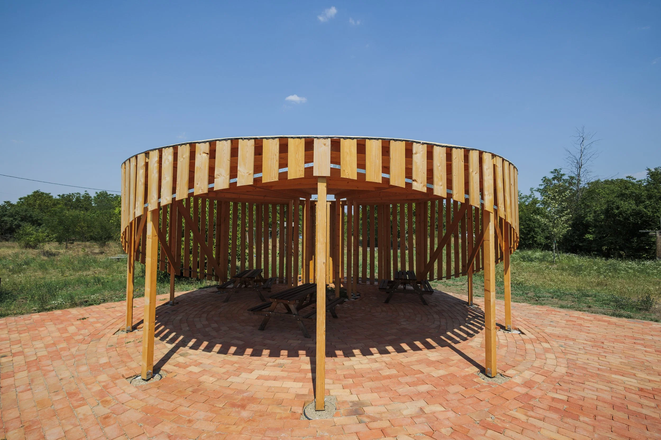 A circular wooden pavilion with a brick floor, surrounded by greenery and trees, under a clear blue sky.