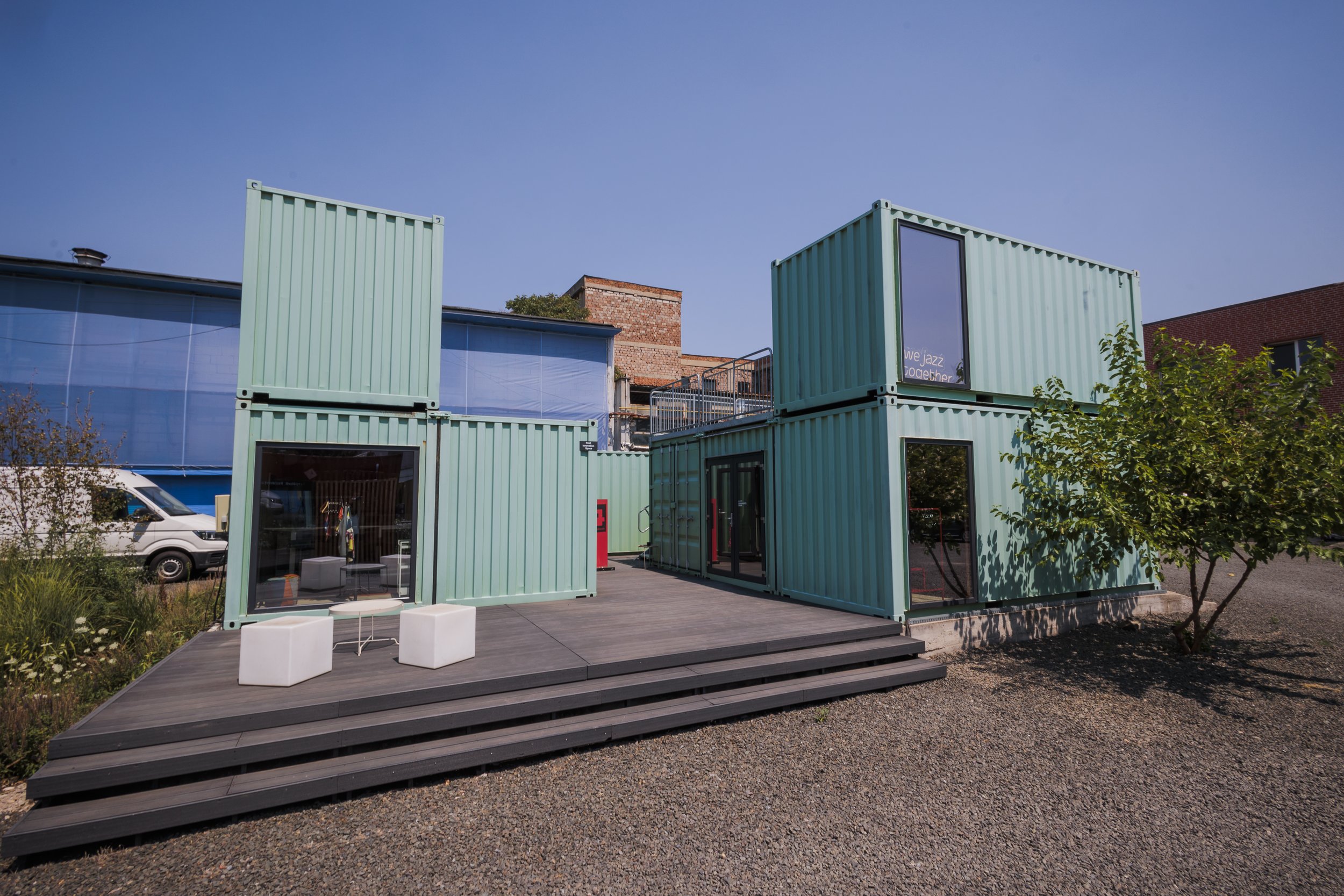 A modern outdoor space made of stacked shipping containers with large windows and a small wooden deck. There are two white chairs and a small table on the deck, with trees and buildings visible in the background.