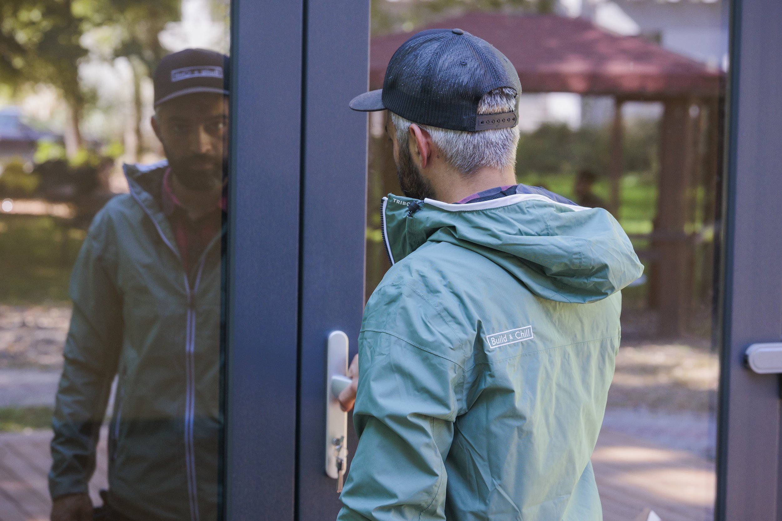 A man with gray hair and a dark baseball cap looks at his reflection in a glass door, wearing a light blue jacket with the words 'Build & Chill' on the back, outdoors with trees in the background.
