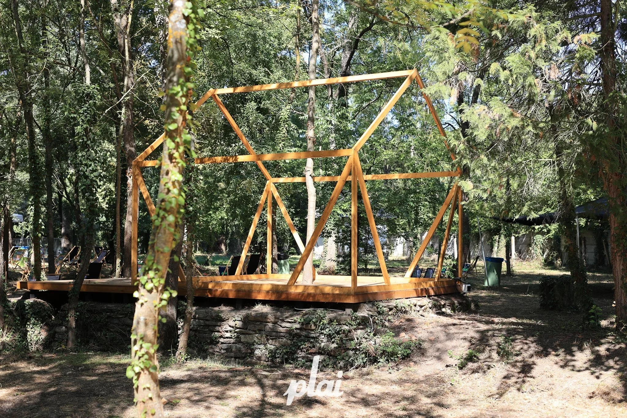 A wooden frame structure under construction in a wooded outdoor area, with trees and sunlight surrounding it.