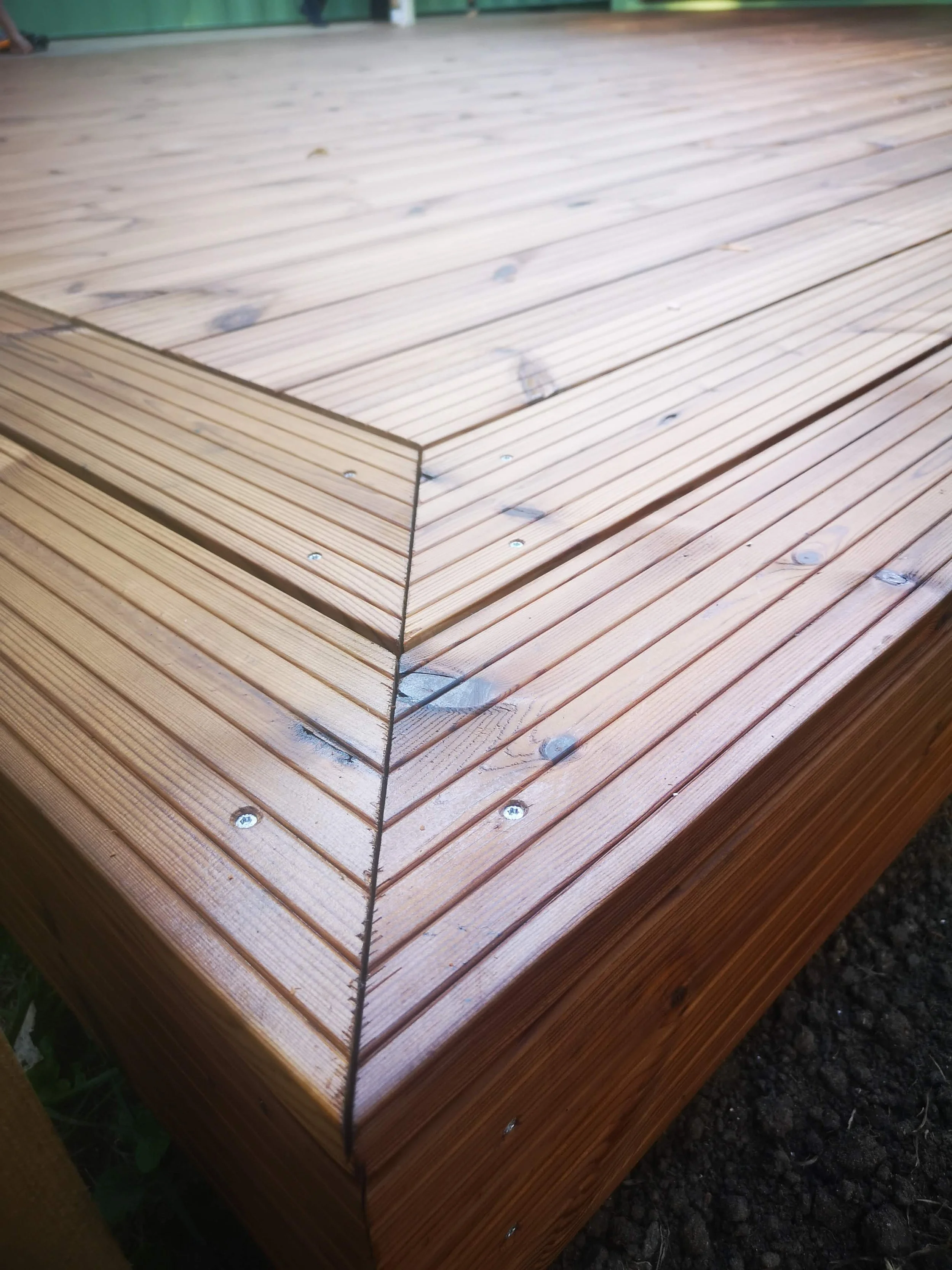 Close-up view of a wooden deck or platform made of horizontally aligned planks, with metal screws securing the planks, and a green background suggesting outdoor setting.