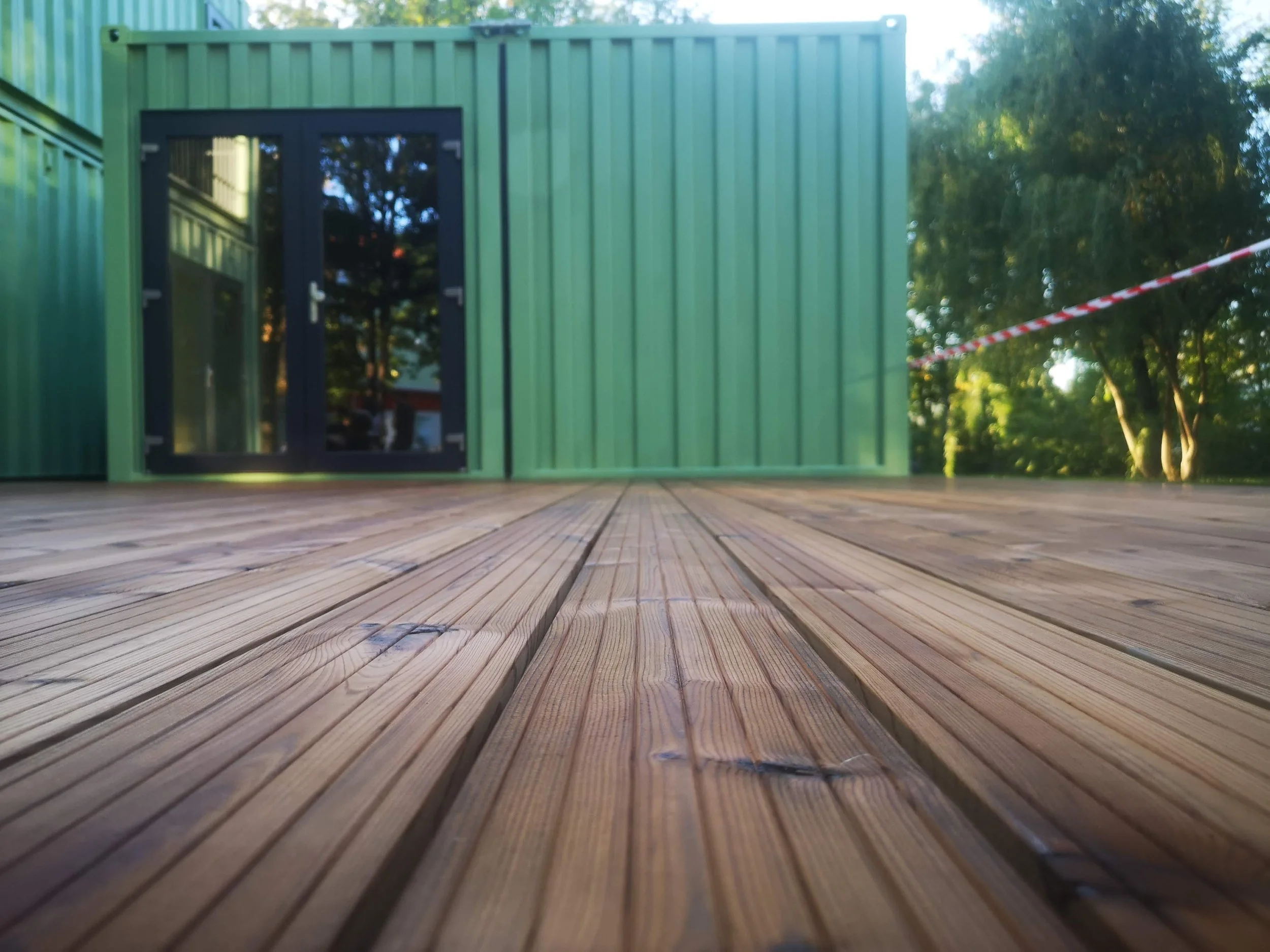 A green shipping container with a black door on a wooden deck, surrounded by trees and greenery during daytime.